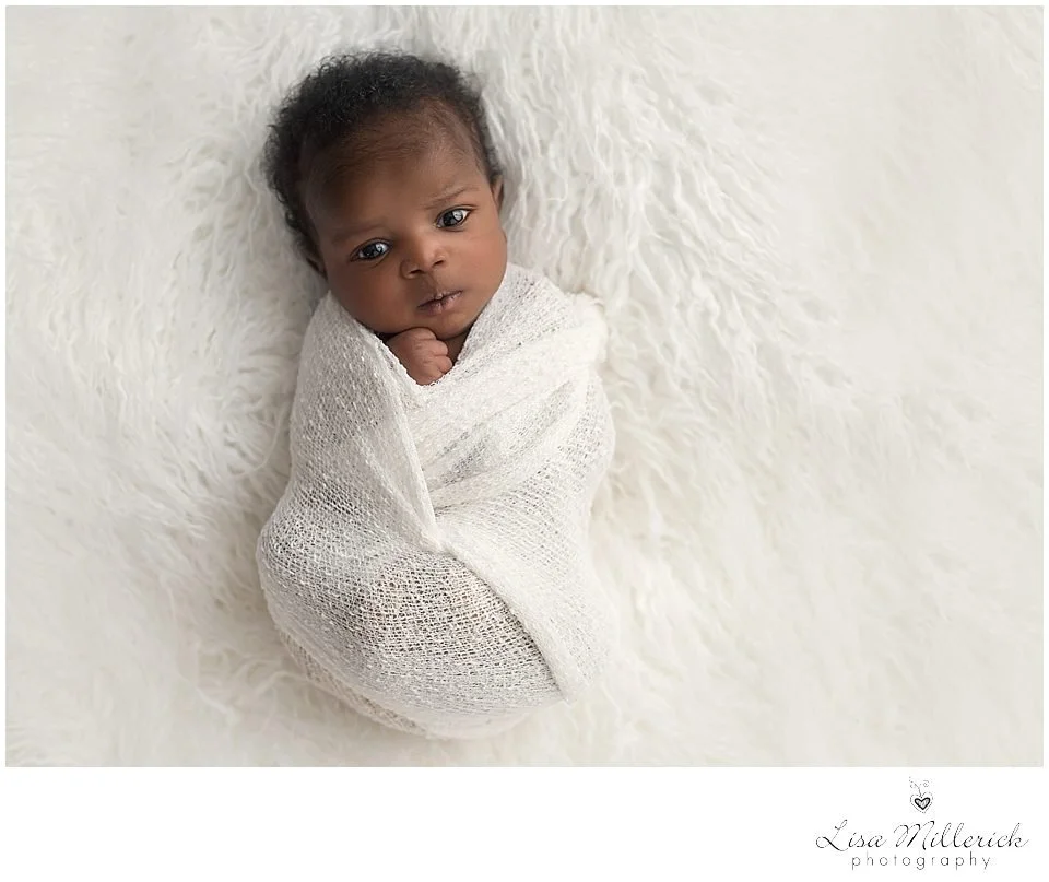 A baby wrapped in a white knit blanket, lying on a fluffy white surface, looking at the camera.