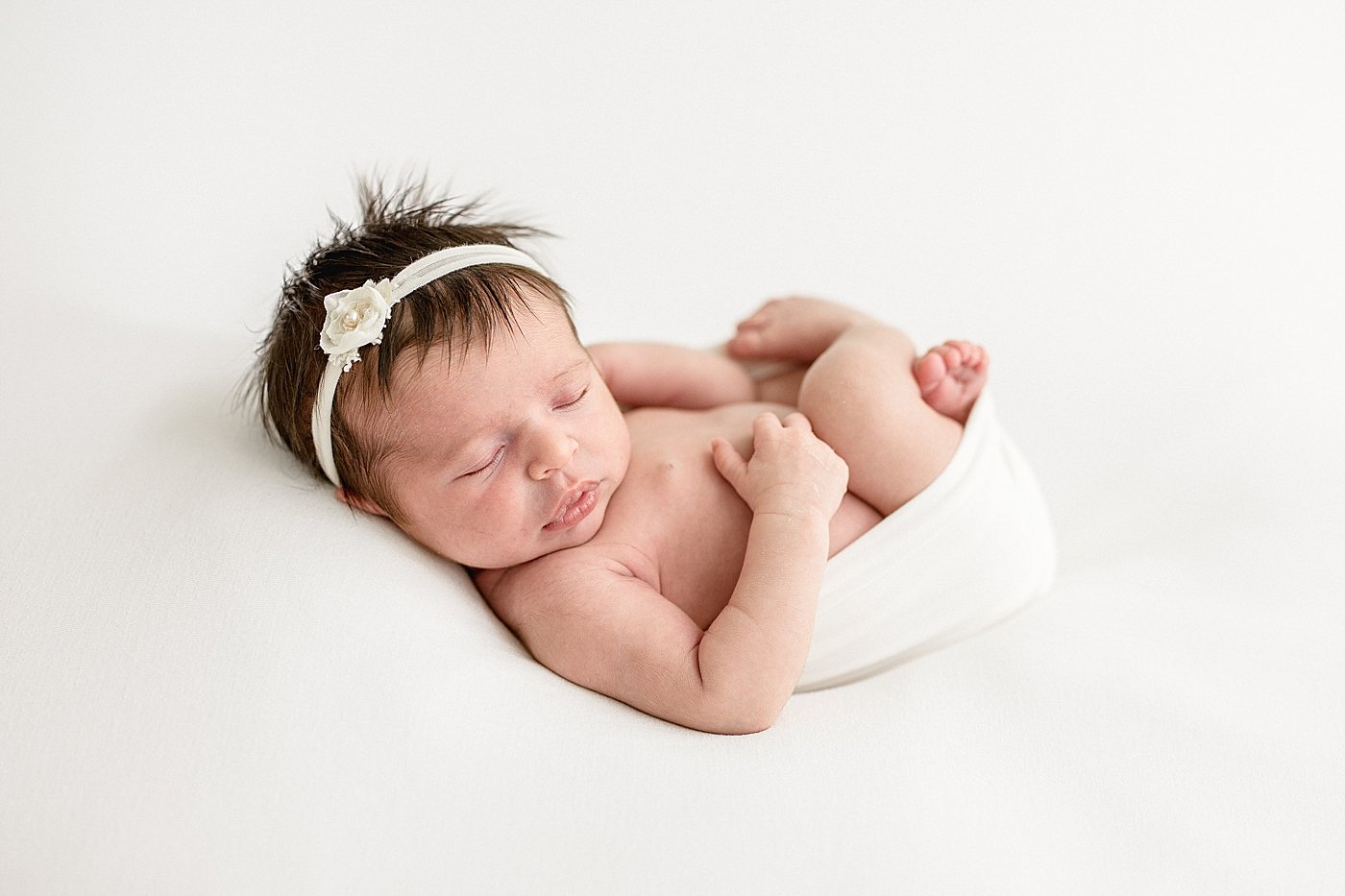 A sleeping newborn baby girl with a white headband and flower, lying on her side on a white blanket.