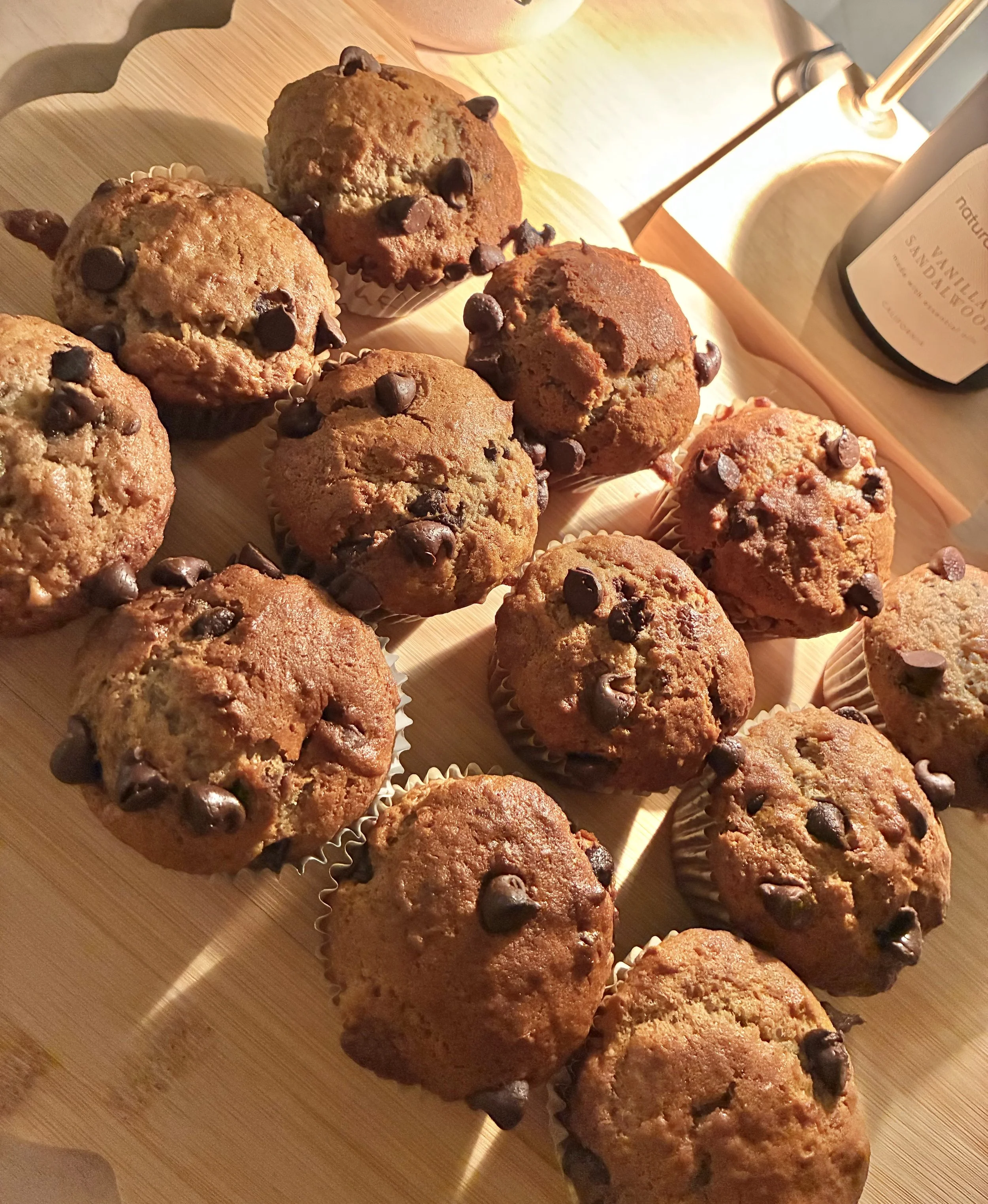 A baking sheet with chocolate chip muffins on a wooden surface, illuminated by warm lighting.