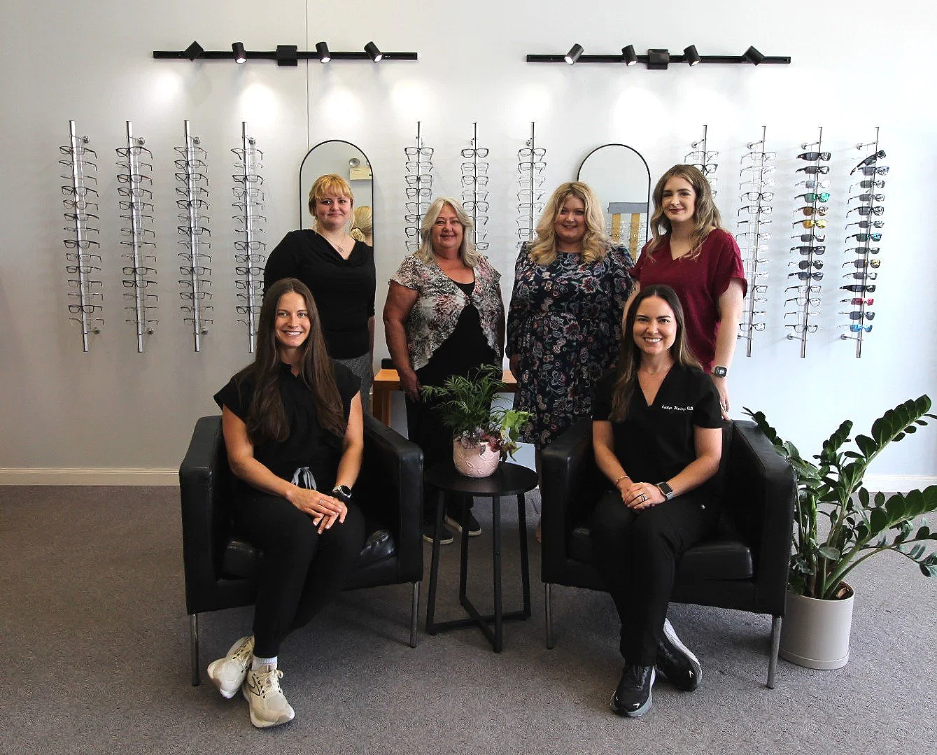Group of six women posing in an eye care or optometry office, with glasses displayed on wall behind them and two black armchairs in front of a small table with a potted plant.