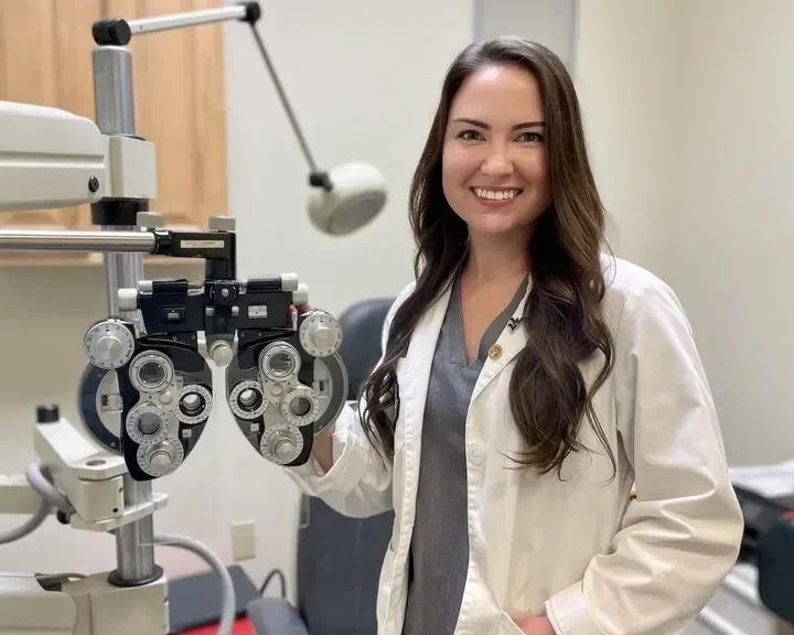 A female eye doctor, dressed in a white coat, standing next to an ophthalmic examination instrument in a medical office, smiling at the camera.