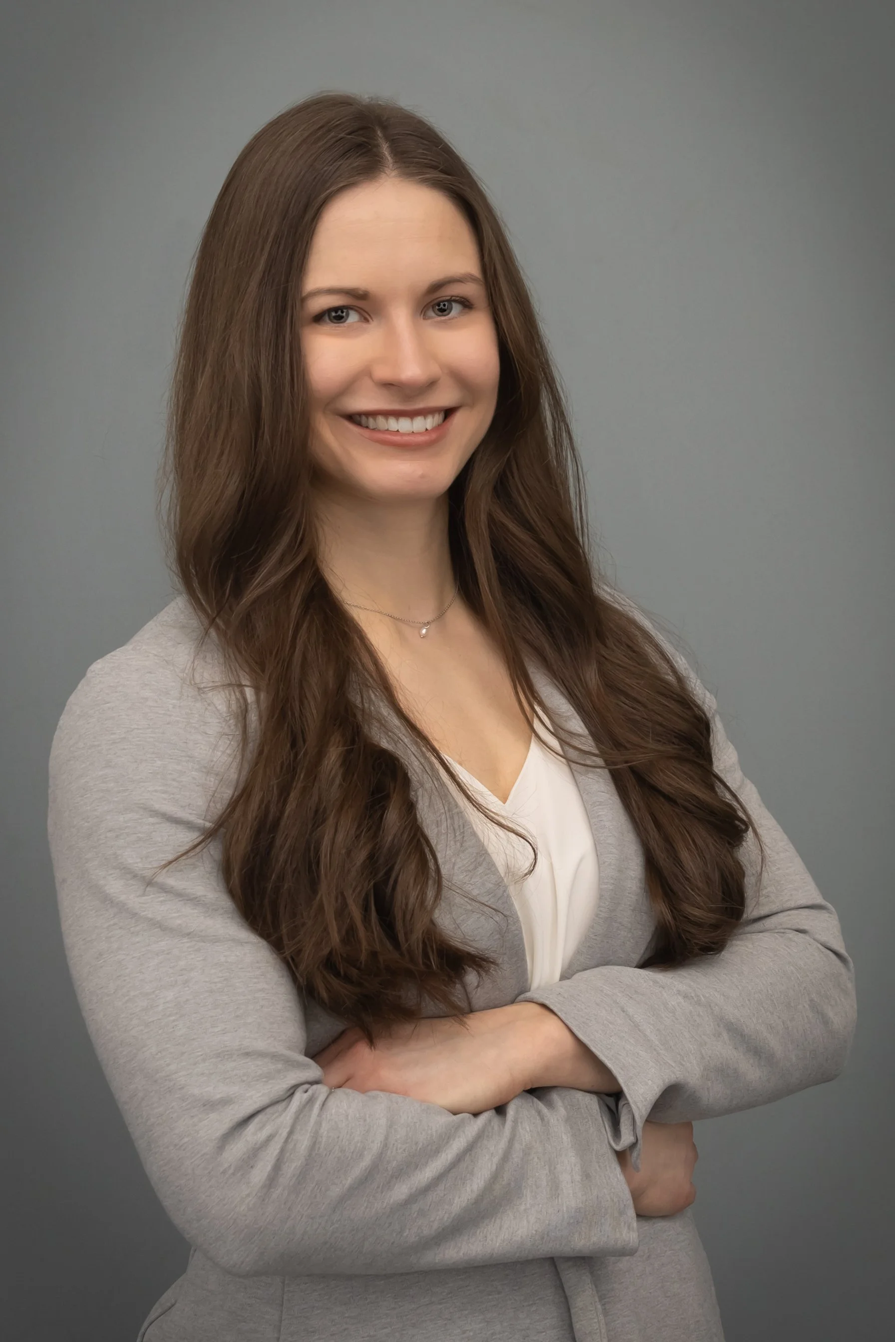 Portrait of a young woman with long brown hair, smiling, wearing a light gray blazer over a white blouse, against a gray background.