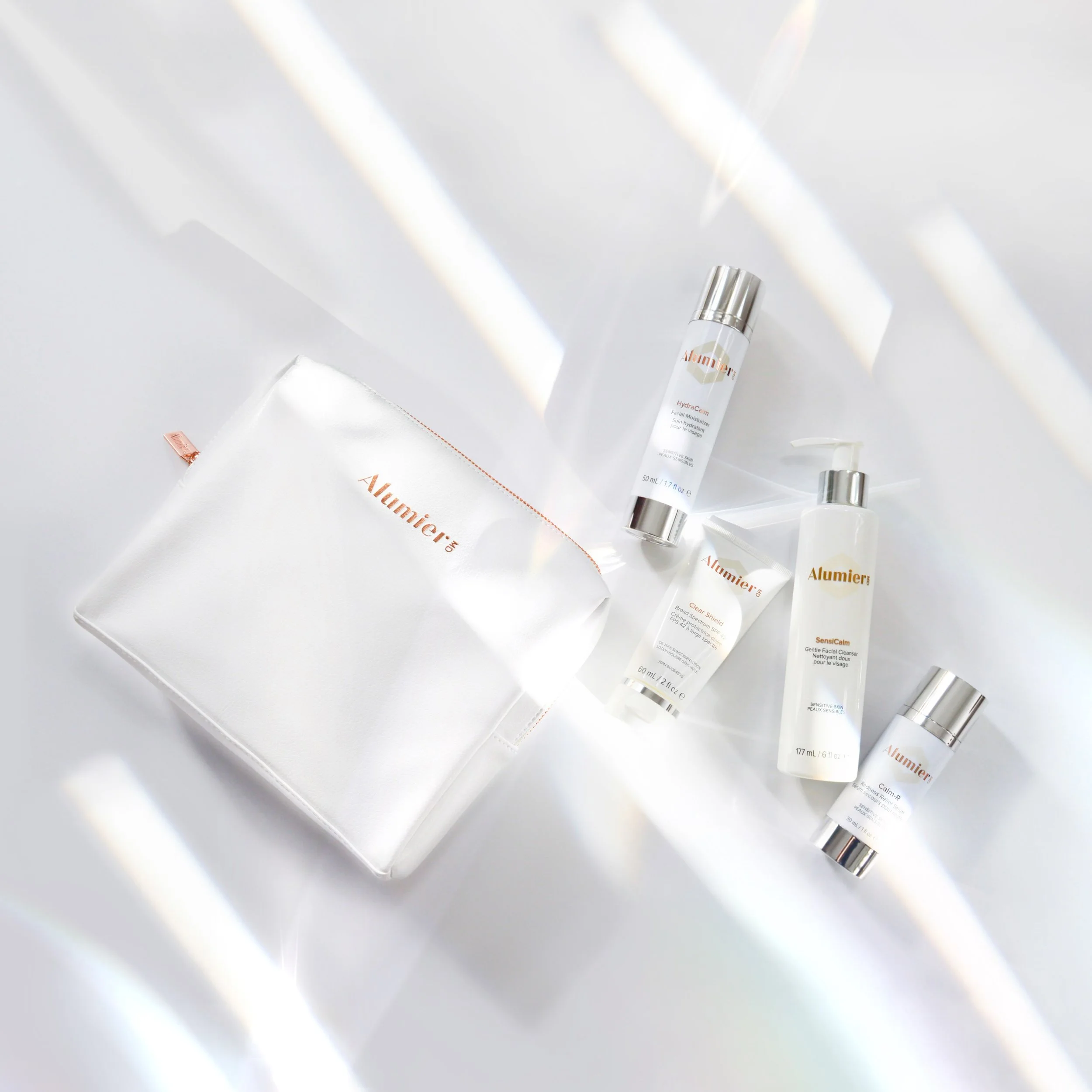 A white cosmetic pouch with the brand name 'Alumier' in orange is lying on a white surface. Beside it are four skincare bottles and tubes, all labeled with the same brand, including a facial moisturizer, cleanser, and other skincare products, all with silver caps.