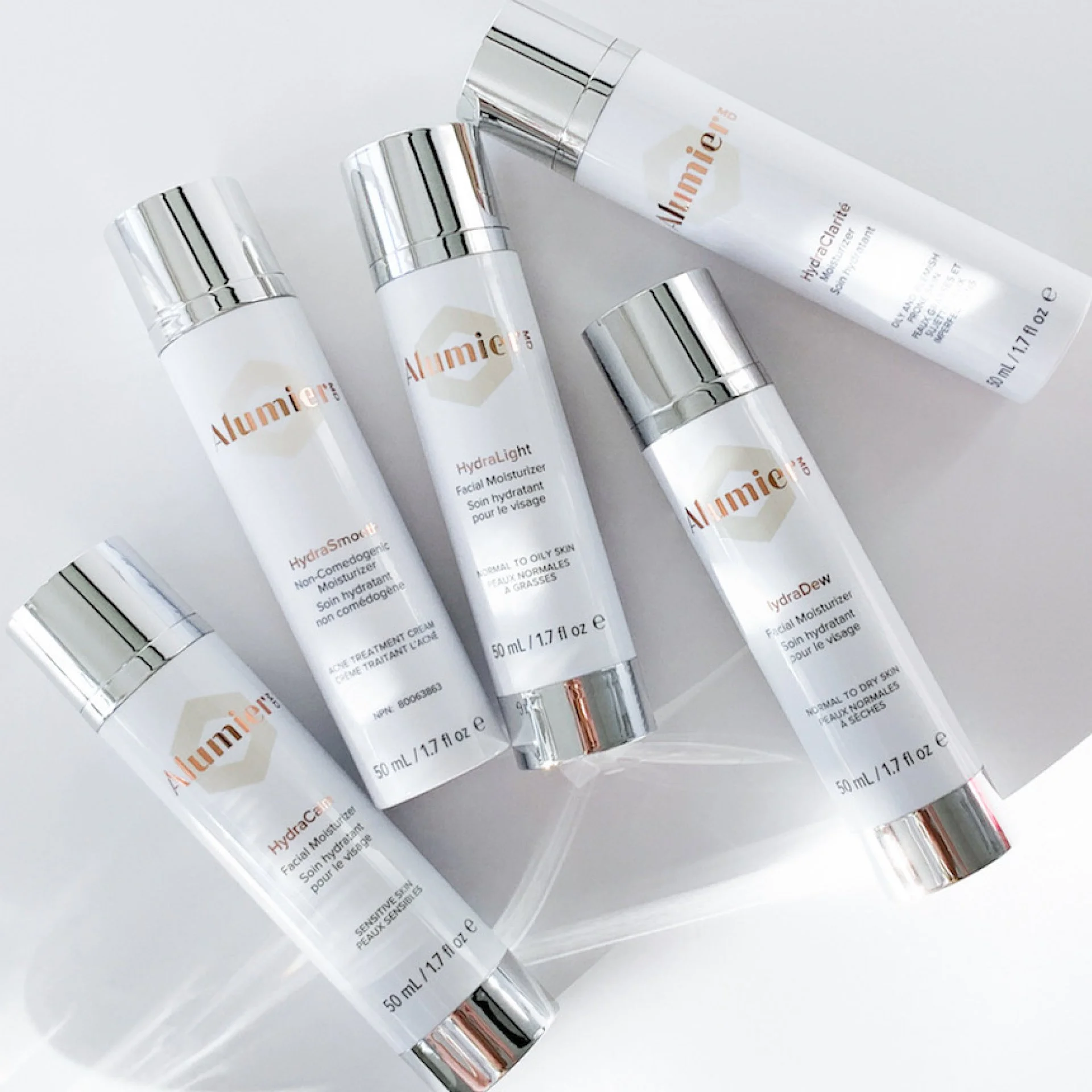 Five bottles of Alumière skincare products, including HydraSmooth, HydraLight, and HydraDew, arranged on a white surface with shadows