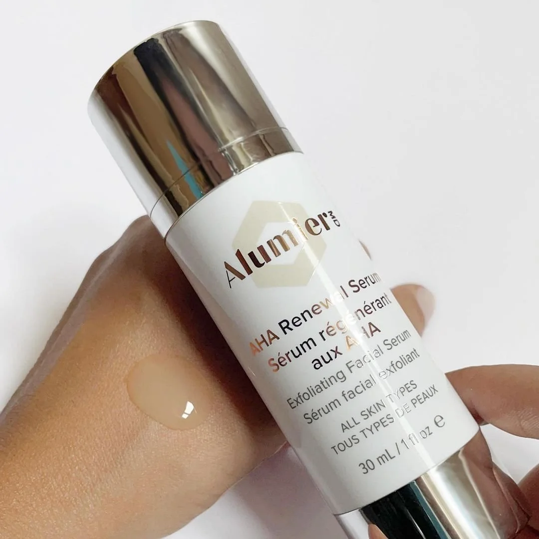 A hand holding a white bottle of AlumierMD AHA Renewal Facial Serum with a silver cap, showing a drop of serum on the back of the hand.