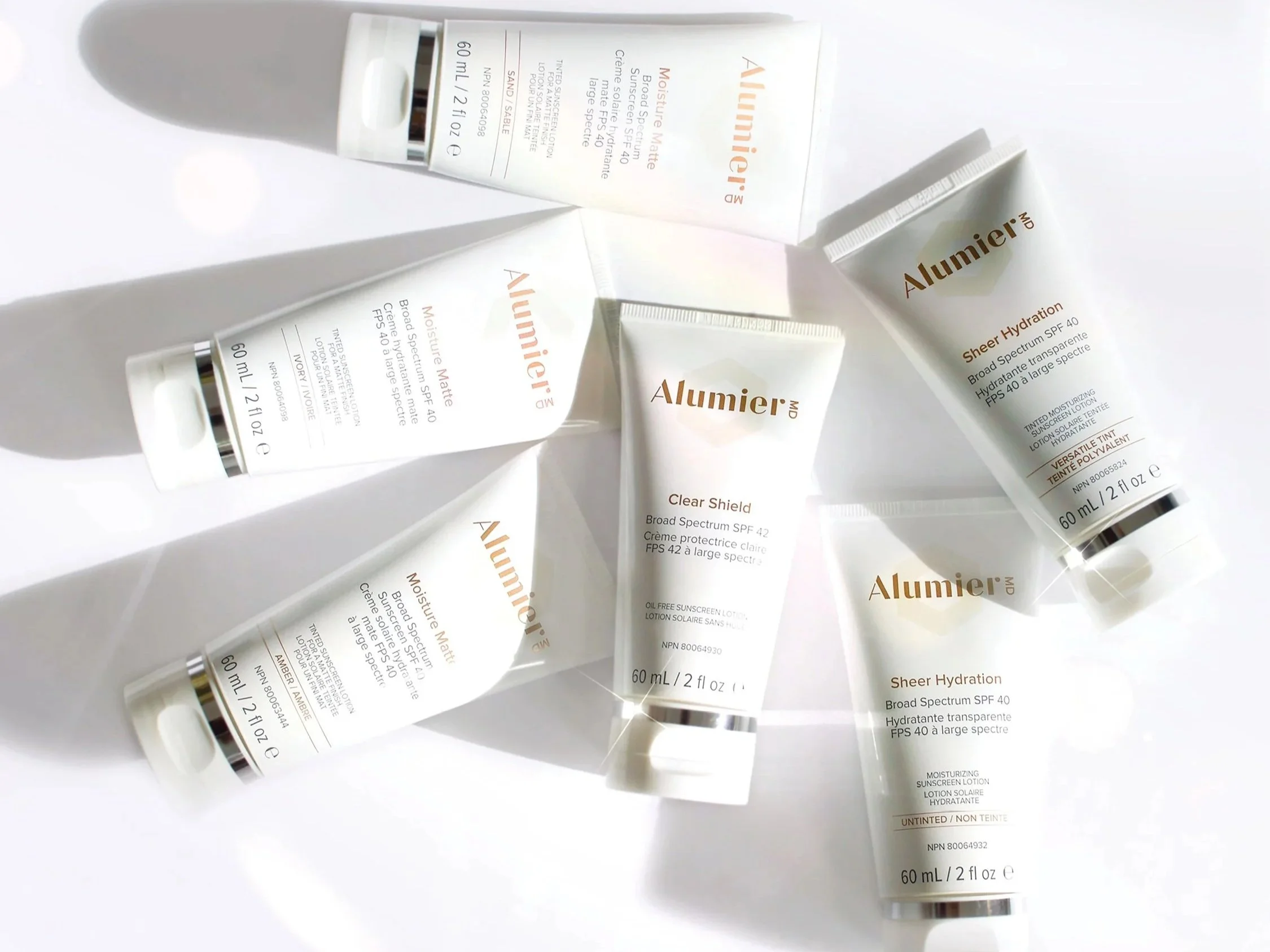 Several skincare products from the brand Alumier, including Moisture Matte, Clear Shield, and Sheer Hydration, arranged on a white surface with shadows cast by the packaging.