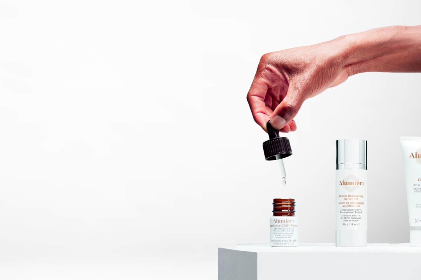 A person's hand holding a dropper, dispensing serum into a small brown bottle labeled 'Alumier', with other skincare products nearby on a white surface against a plain light gray background.