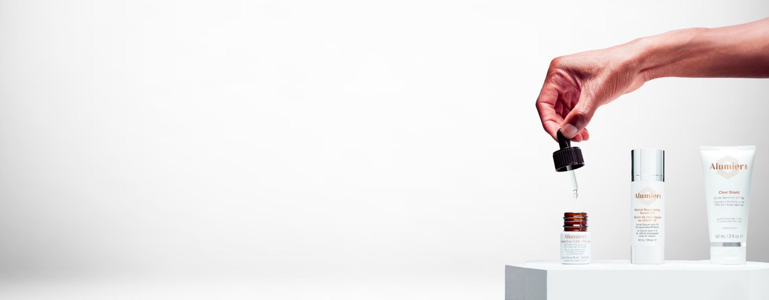 Hand holding a dropper above a small jar of skincare product with two more skincare products beside it on a white surface, against a plain white background.