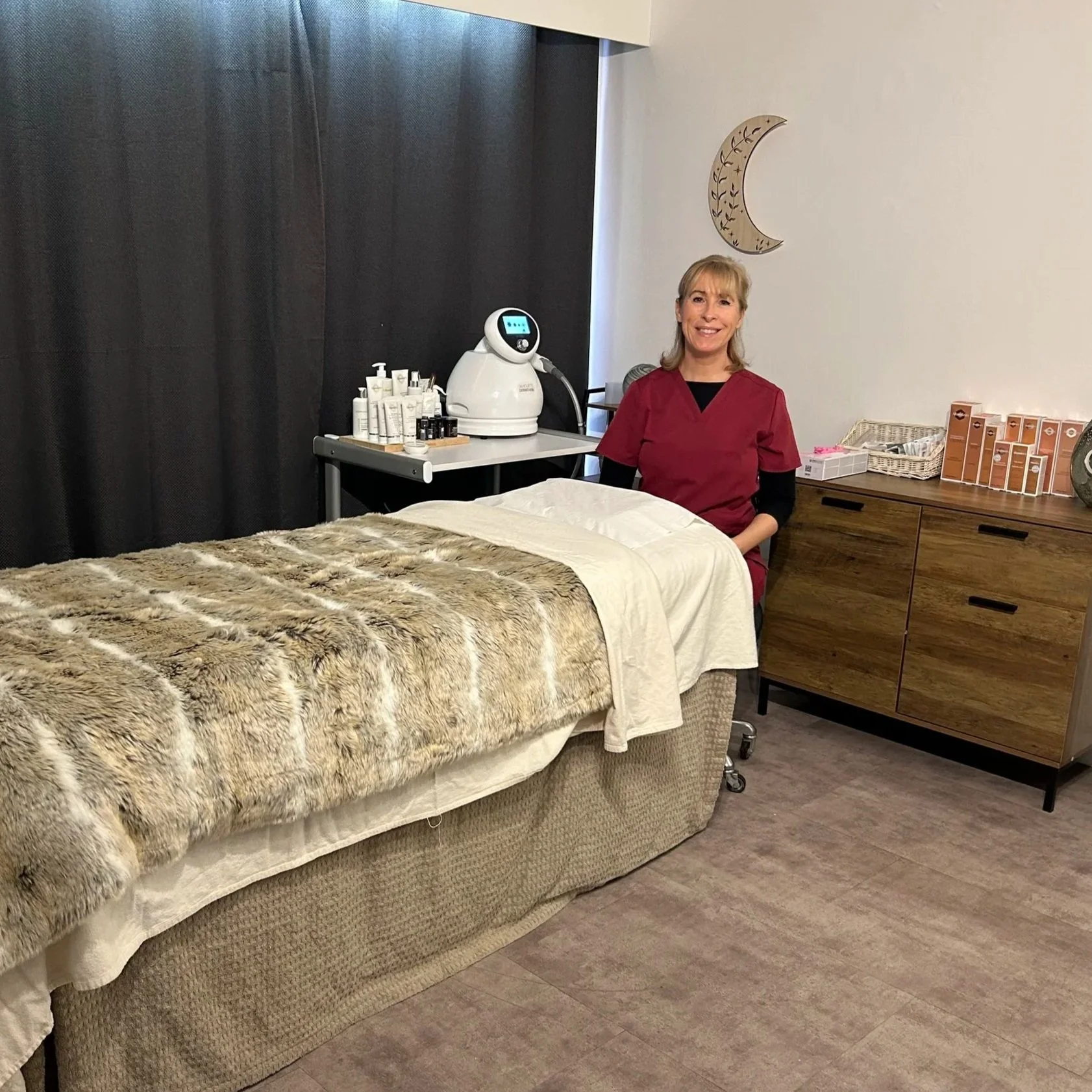 A woman in red scrubs sitting at the edge of a massage table in a spa or clinic room, with a fur blanket on the table and skincare products and equipment on a rolling cart and a wooden cabinet beside her.