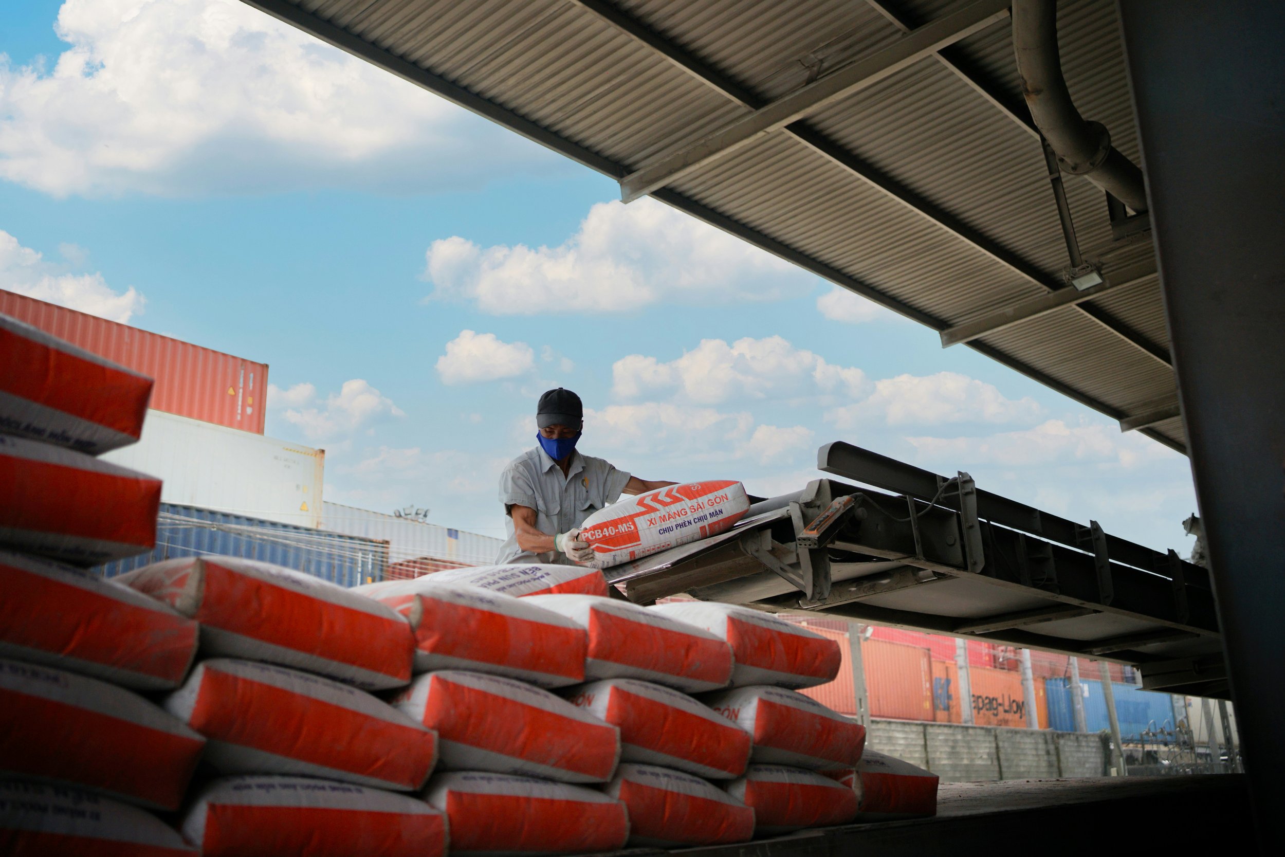 A worker wearing a blue face mask and gloves loads bags of cement onto a truck at a shipping yard, with cargo containers and a cloudy sky in the background.