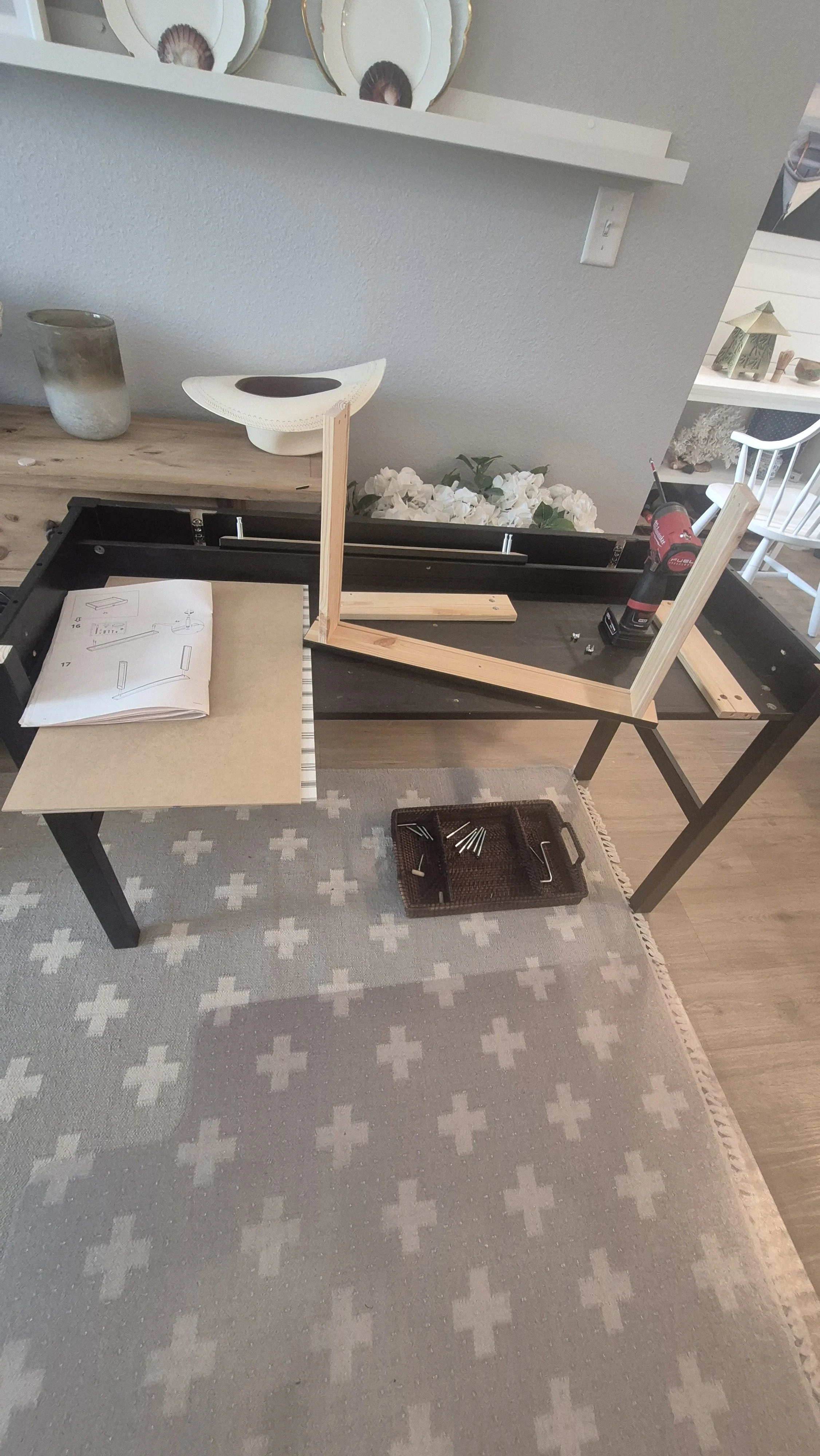 A workspace with a black table set up for assembling furniture, including wooden pieces, an instruction manual, a power drill, and screws. Decorative dishes and flowers are visible on a shelf and table in the background; Golden, Colorado