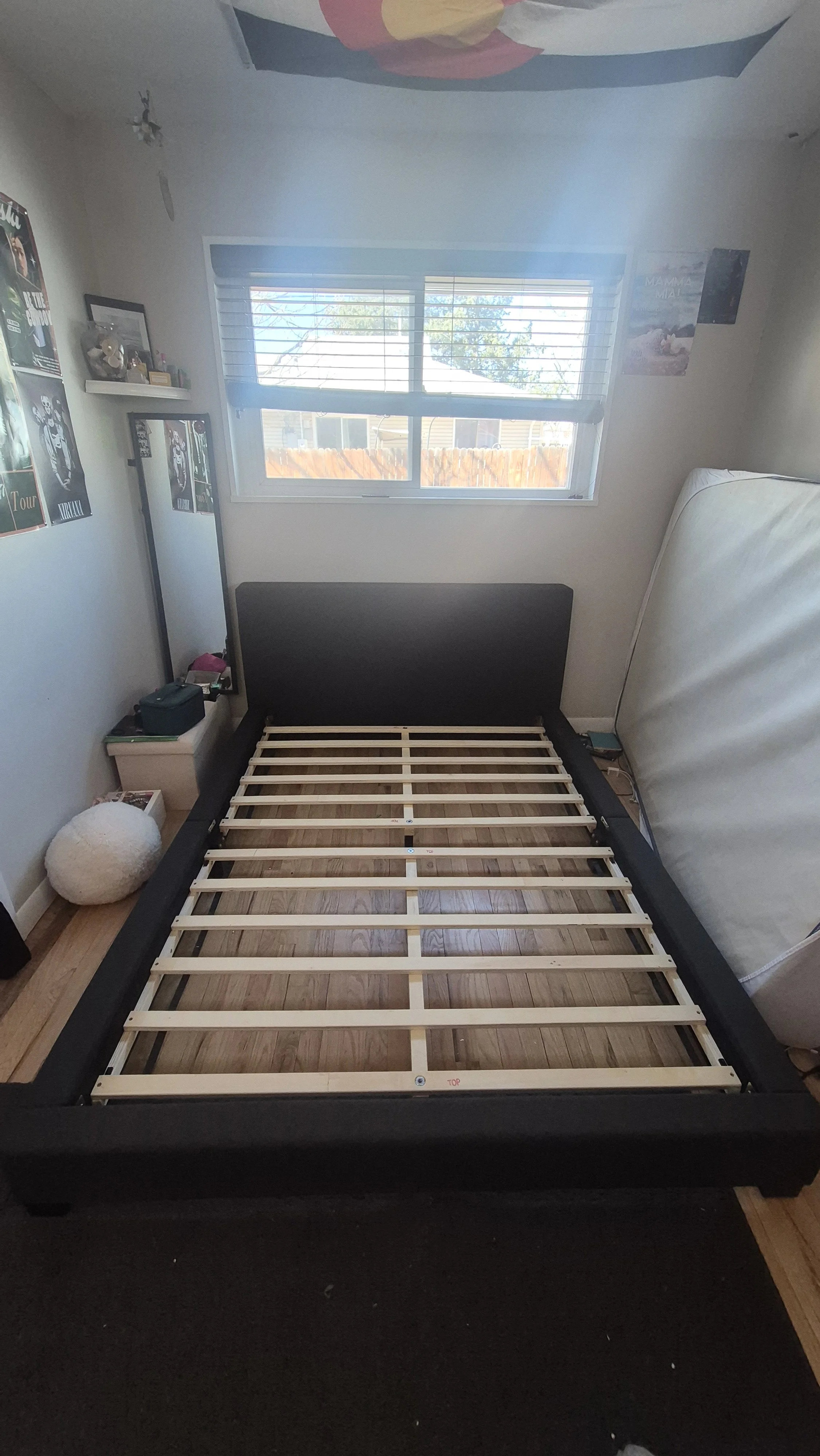 Unassembled bed frame in a bedroom with a window, posters on the wall, a mirror, a white fuzzy ball, and various personal items on a nightstand; Westminster, Colorado