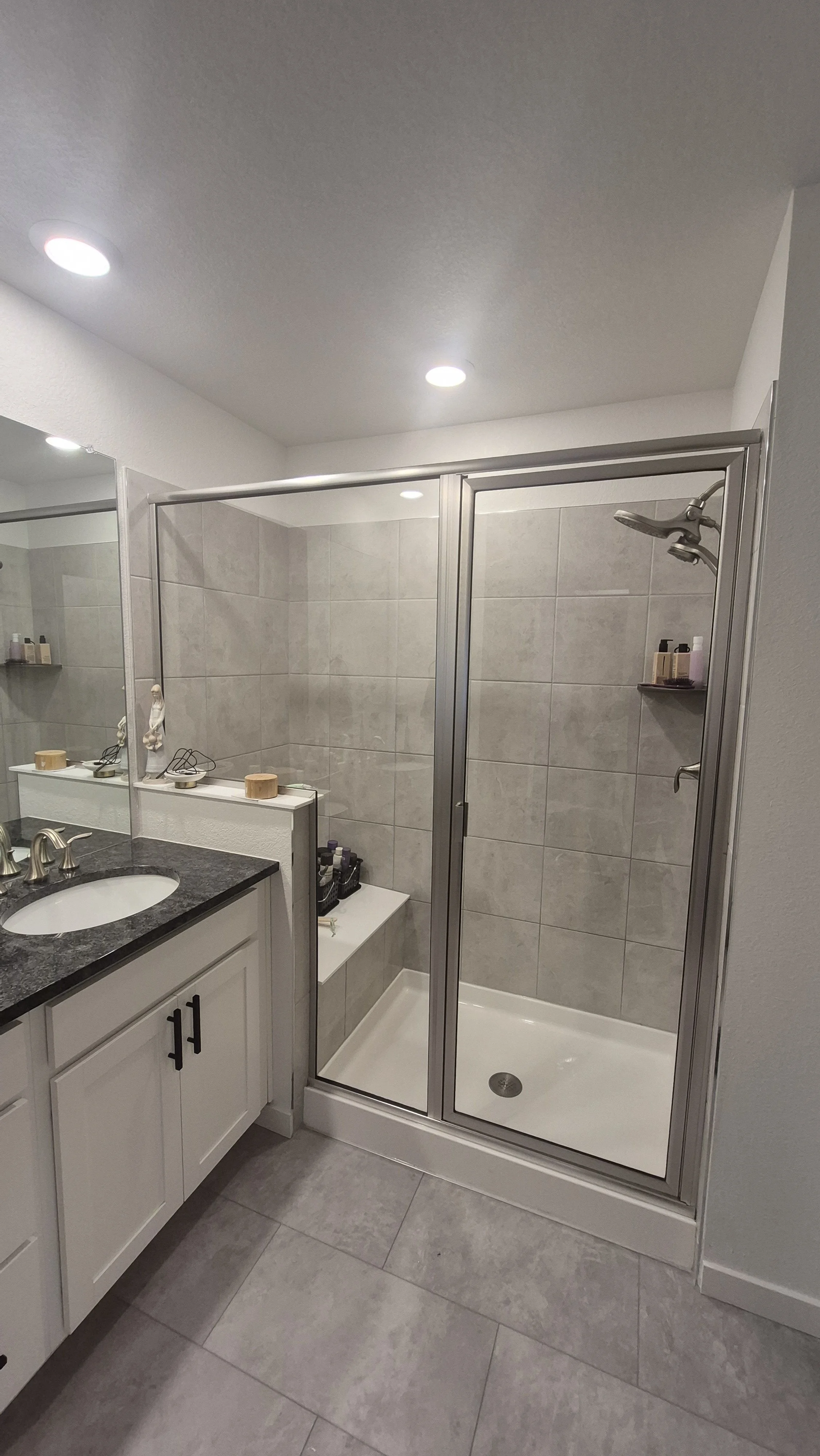 A bathroom with a shower, a white vanity with a black countertop, and various toiletries and decorative items on the shelves and countertop.
