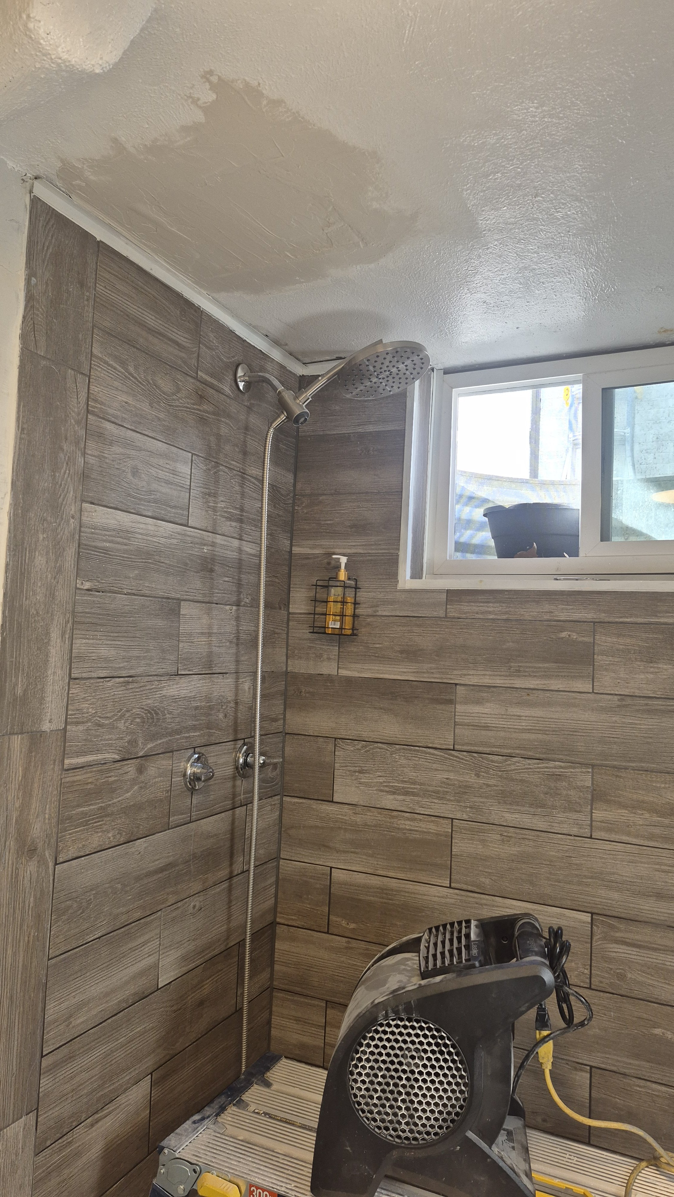 A bathroom shower area with brown wood-textured tiles, a showerhead, and a window, with a heater or fan device in the foreground; Arvada, Colorado