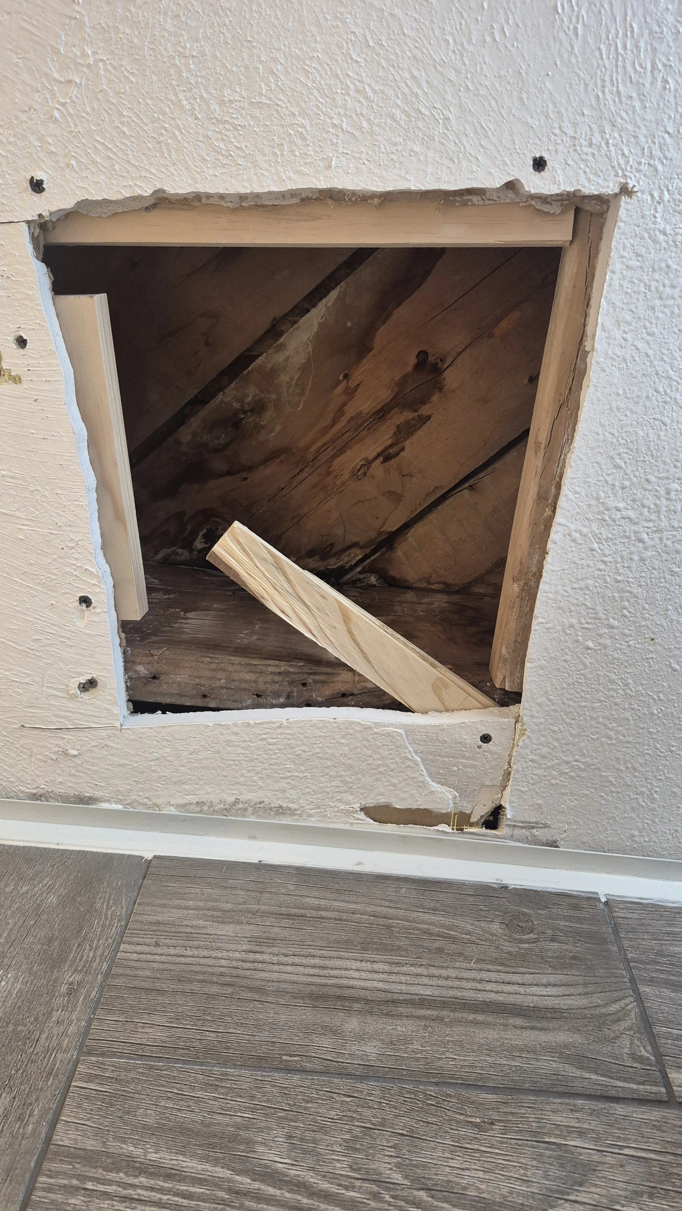 Ceiling with a square hole revealing the attic space with wood and insulation, partially boarded up with an angled wooden piece. Exterior wall with textured paint and wooden flooring; Central Park, Denver, Colorado