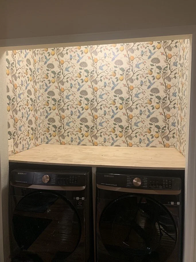 Laundry room with wallpaper featuring birds, trees, and fruits, with a wooden platform above two black Samsung washing machines.