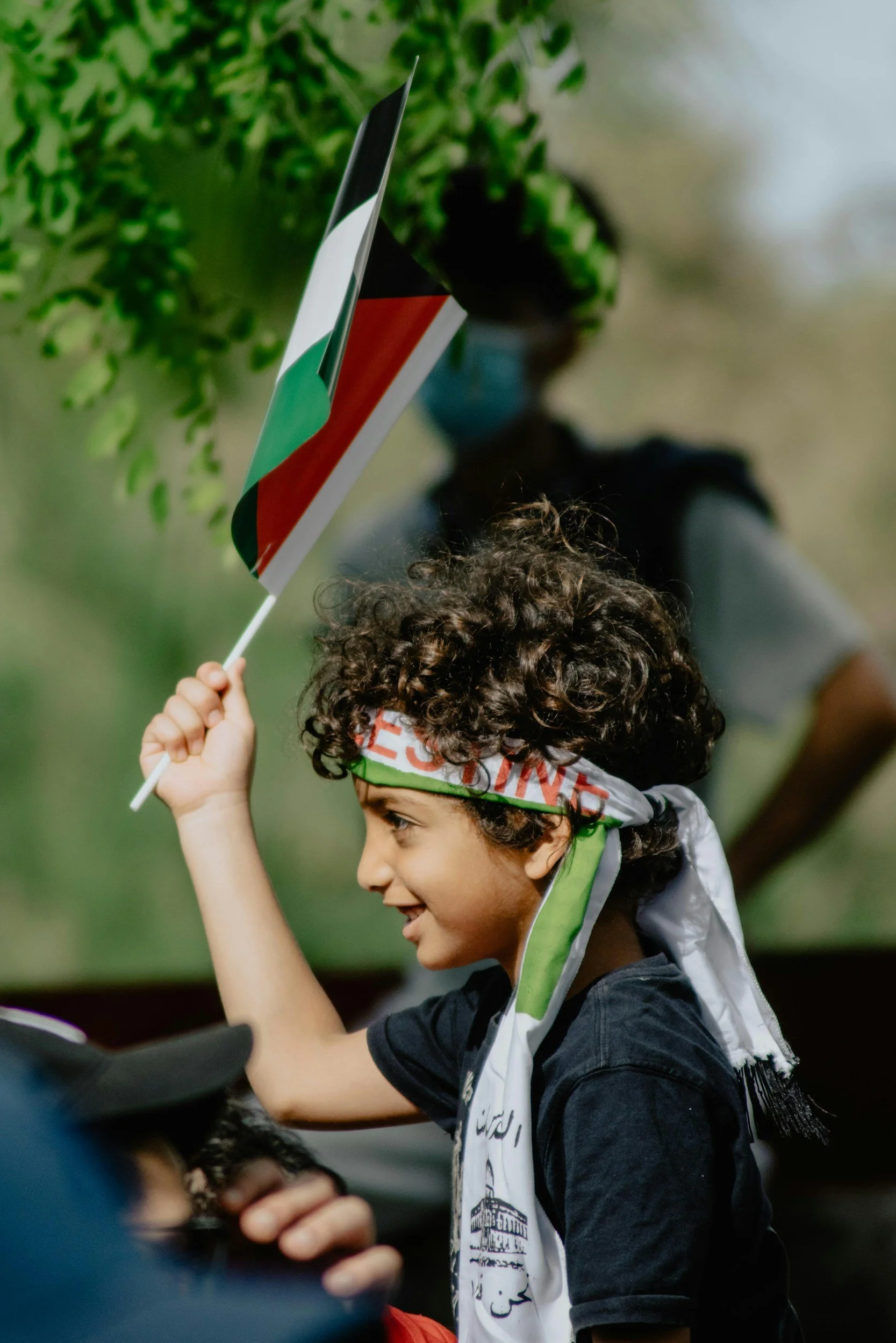 A young boy with curly hair and a headband is waving a small Palestinian flag. He is smiling and appears to be at a rally or demonstration, with a person in the background wearing a face mask and standing outdoors.