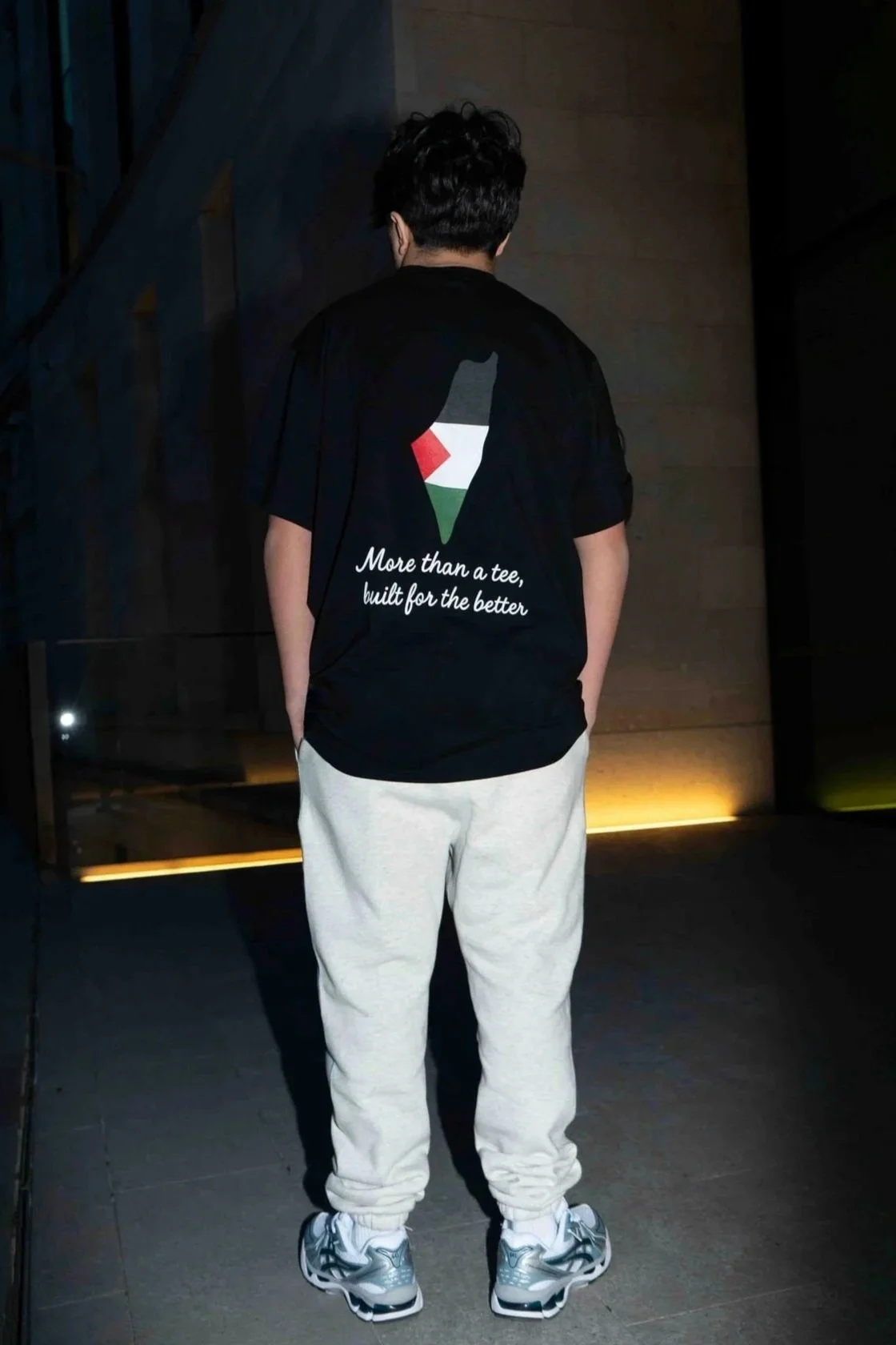 Person standing with their back to the camera, wearing a black T-shirt with a graphic and white text on the back, light-colored sweatpants, and sneakers, in a dimly lit indoor setting.