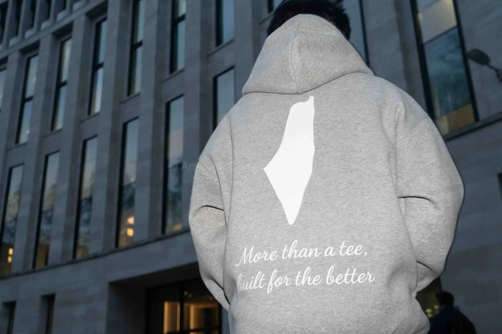 Person wearing a gray hoodie with the hood up, facing away, standing outside a modern glass building at dusk. The hoodie has a white graphic of a shark fin on the back with the text, 'More than a tee, built for the better.'