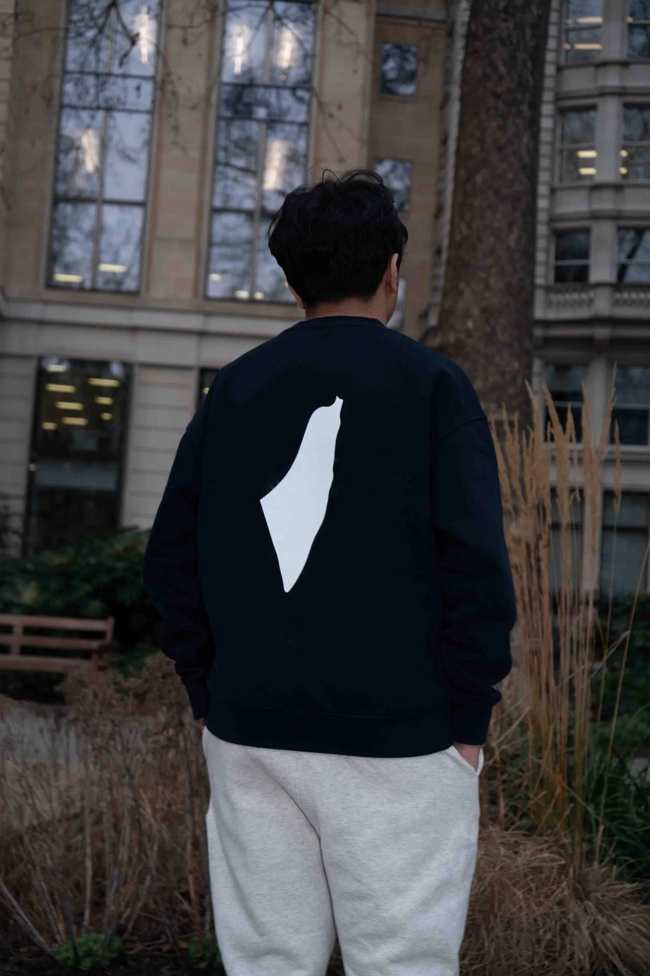 Back of a person wearing a black jacket with a white geometric shape on the back, standing outdoors near a building with large windows and tall trees.