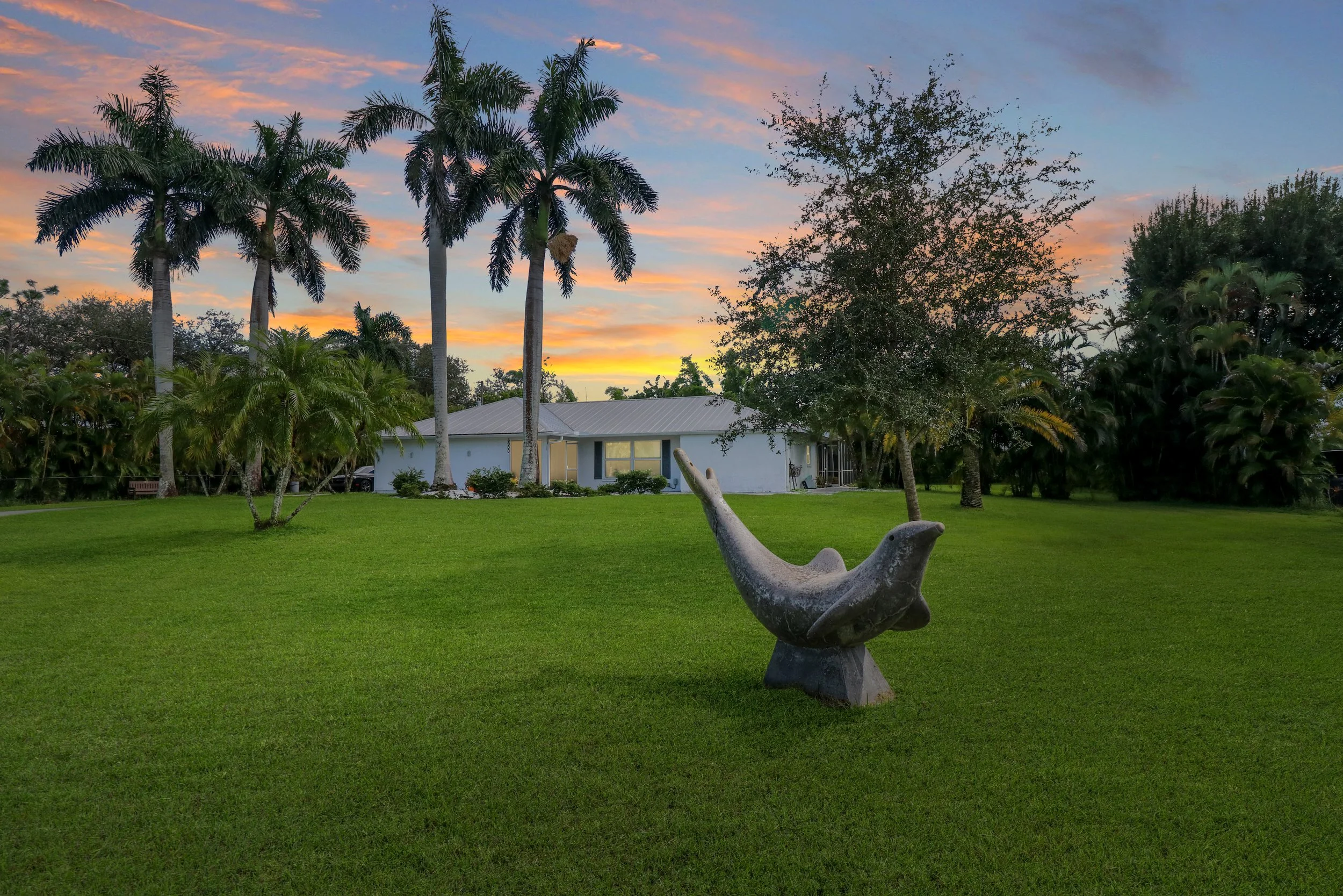 A sculpture of a whale in the foreground on a grassy lawn, with palm trees, a house, and a colorful sunset sky in the background.