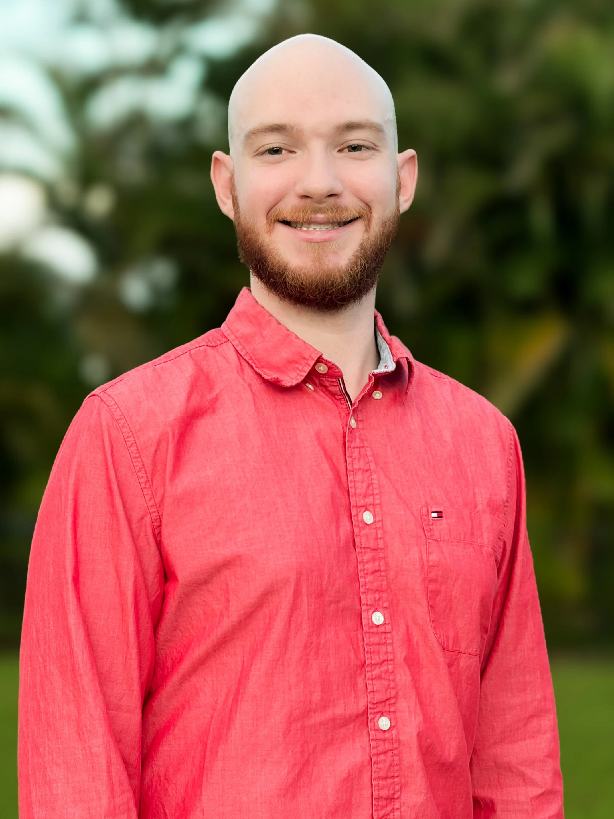 A smiling man with a beard and no hair wearing a red button-up shirt standing outdoors with greenery in the background.
