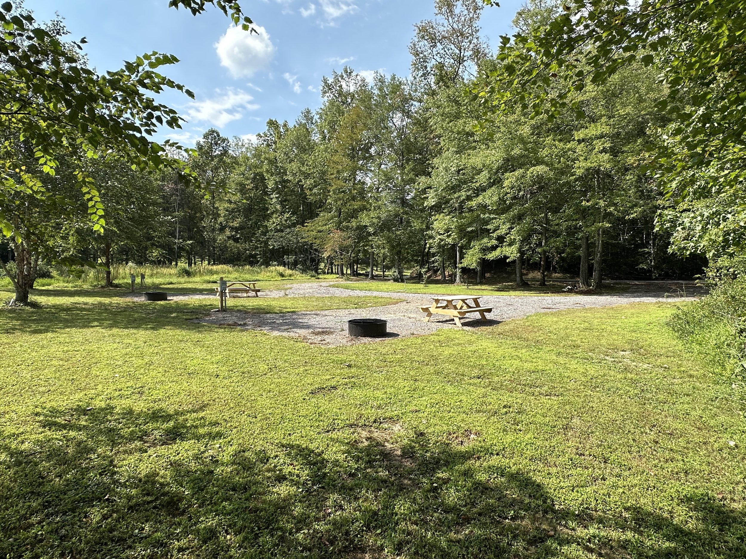 An RV area with a camping or picnic site, featuring two wooden picnic tables, surrounded by trees with green leaves, a gravel pathway, and a few metal fire rings, under a partly cloudy blue sky.