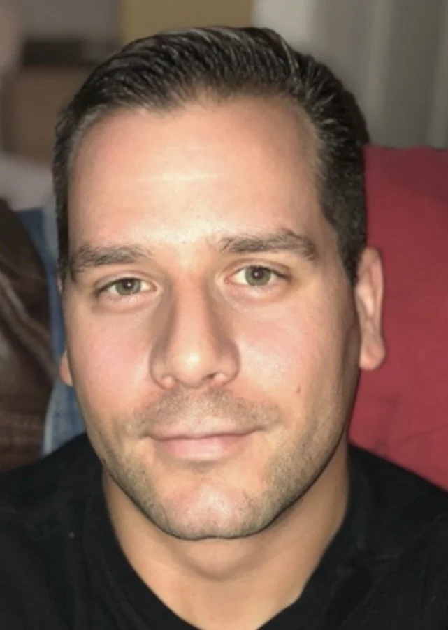 Close-up photo of a man with short dark hair, light skin, and green eyes, smiling slightly, wearing a black shirt.