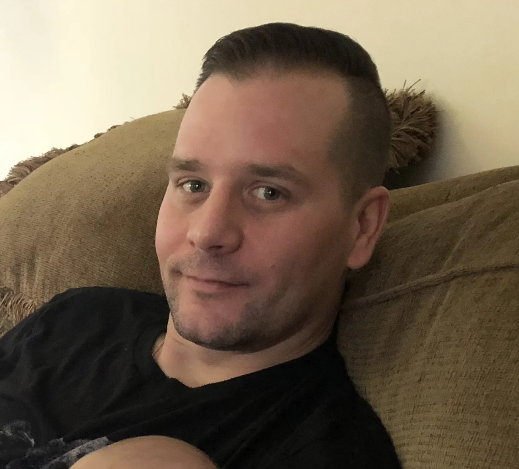 A man with short brown hair, light skin, and a beard, sitting on a brown sofa, looking at the camera.