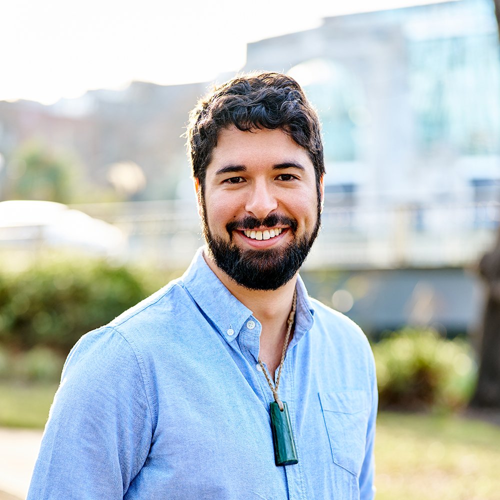 A smiling man with a beard and short dark hair, wearing a light blue button-up shirt with rolled-up sleeves, standing outdoors on a balcony with a blurred green and brown natural background.