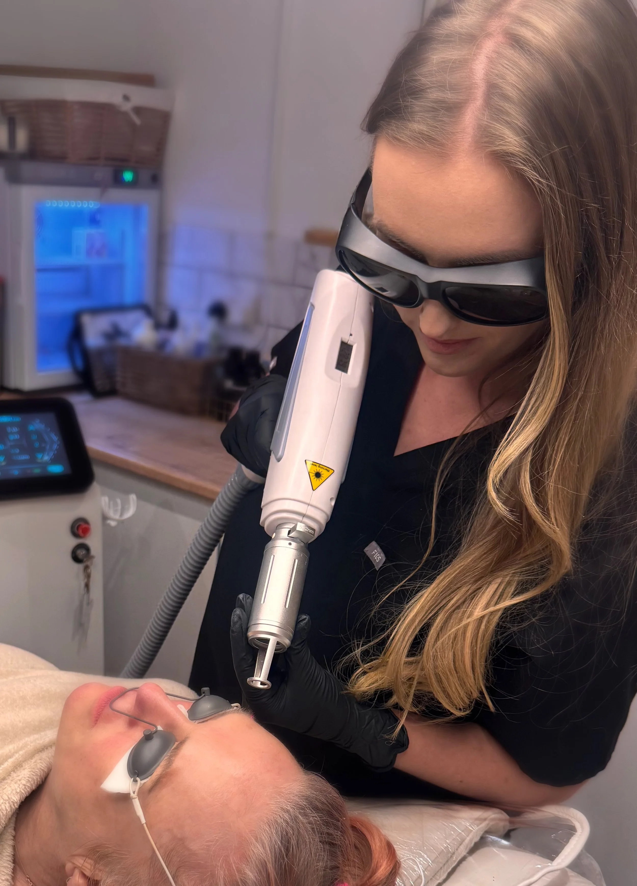 A woman with long blonde hair, wearing black gloves and protective glasses, performs laser skin treatment on a woman lying on a medical bed. The patient has protective eye covers on.