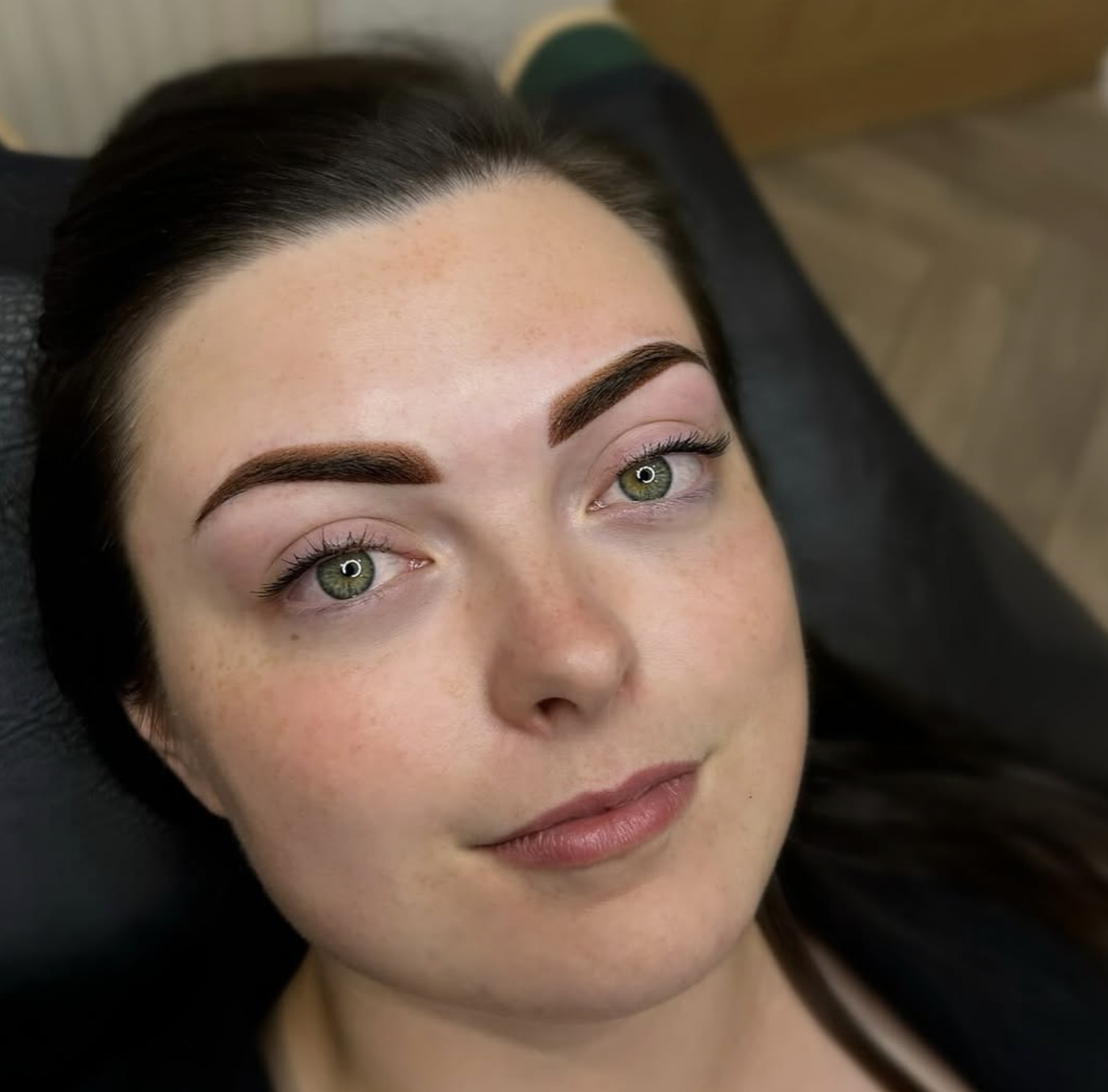 Close-up of a young woman with dark hair, green eyes, and natural makeup, lying back on a black surface.