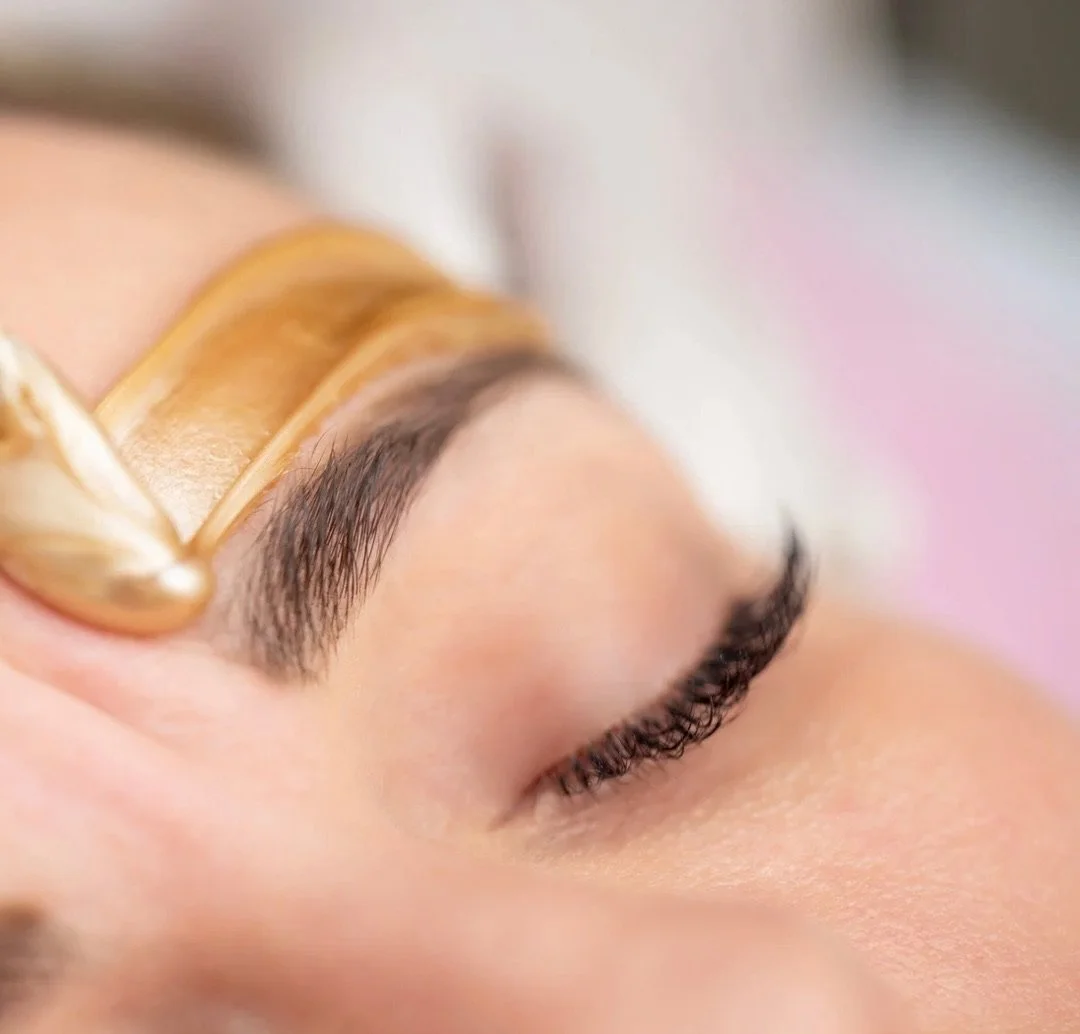 Close-up of a person's closed eye with long eyelashes and groomed eyebrow, with a golden tool applied to the eyebrow.