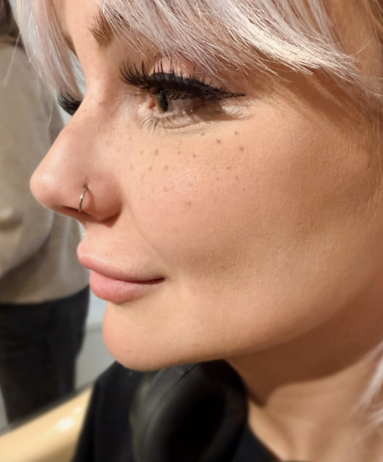 Close-up of a woman's face in profile showing makeup, long eyelashes, freckles, a nose ring, and platinum blonde hair.