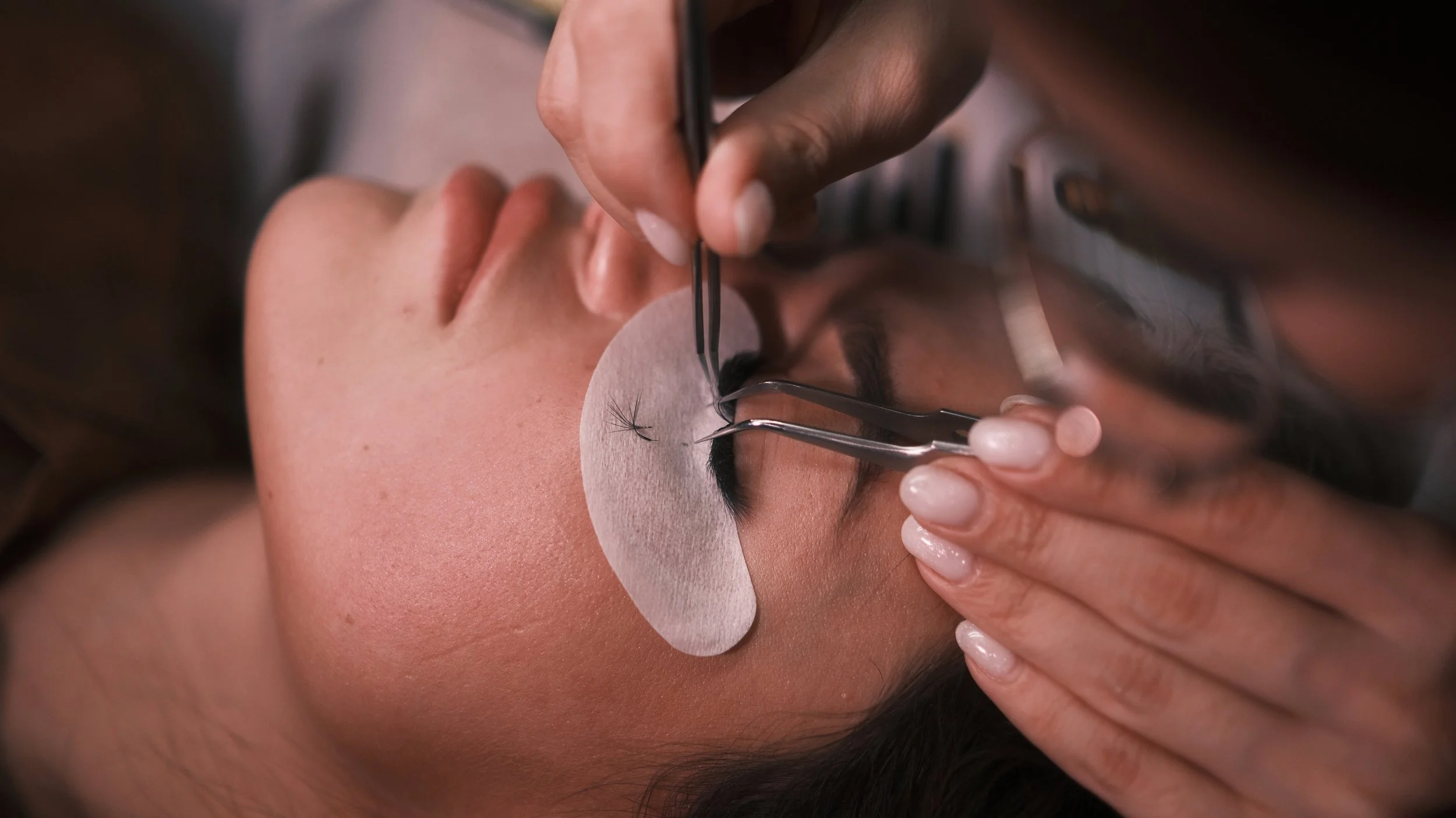 A woman getting eyelash extensions done by a technician using tweezers, with a protective oval pad under her eye.