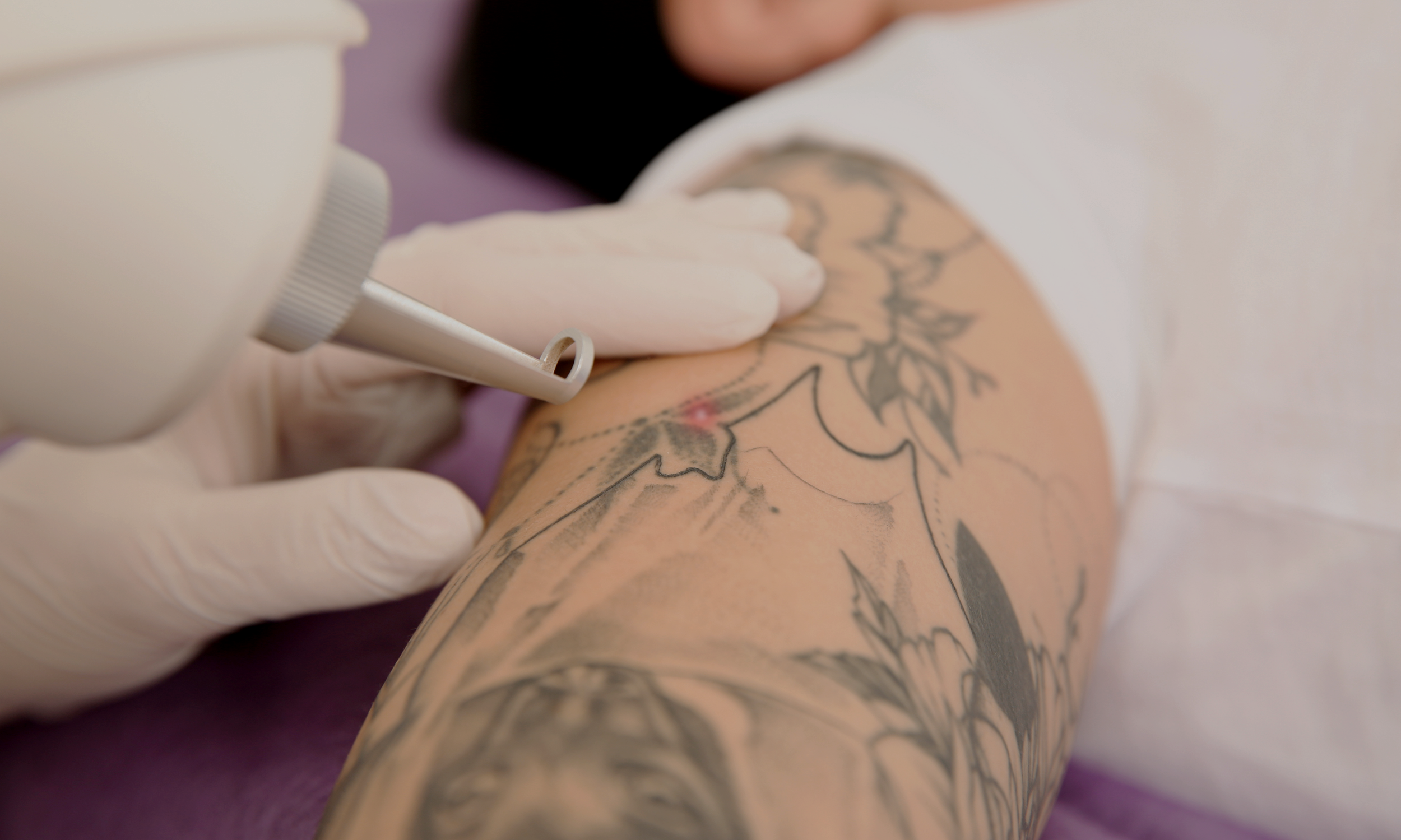 A person in white gloves tattooing a floral design on someone's arm with a tattooing machine.