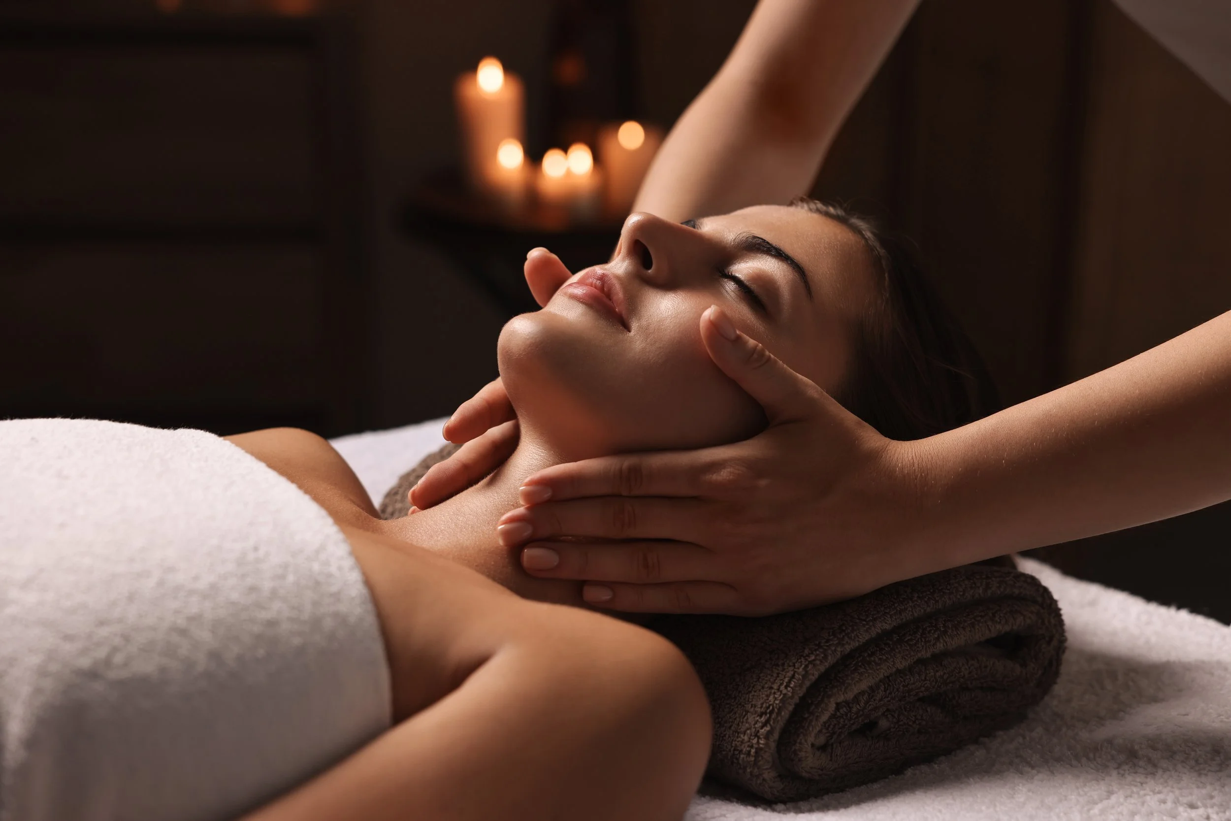 A woman receives a facial massage at a spa, lying on a massage table with her eyes closed, a towel over her chest, and candles in the background creating a relaxing atmosphere.