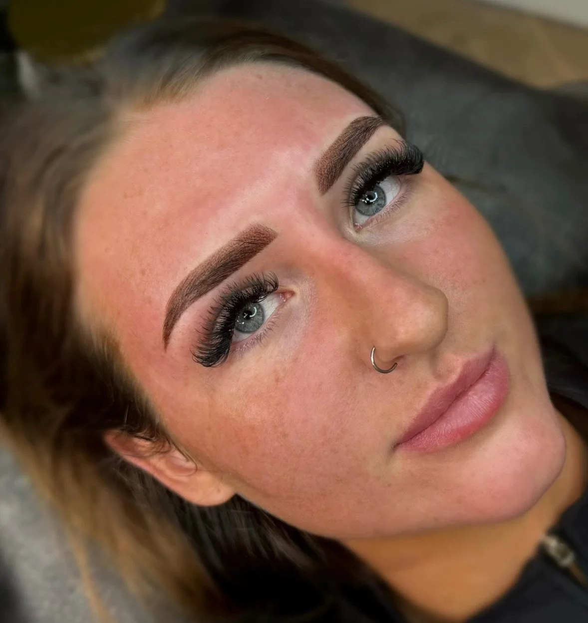 A woman with makeup, including defined eyebrows, long eyelashes, and a nose ring, lying down with her eyes open.