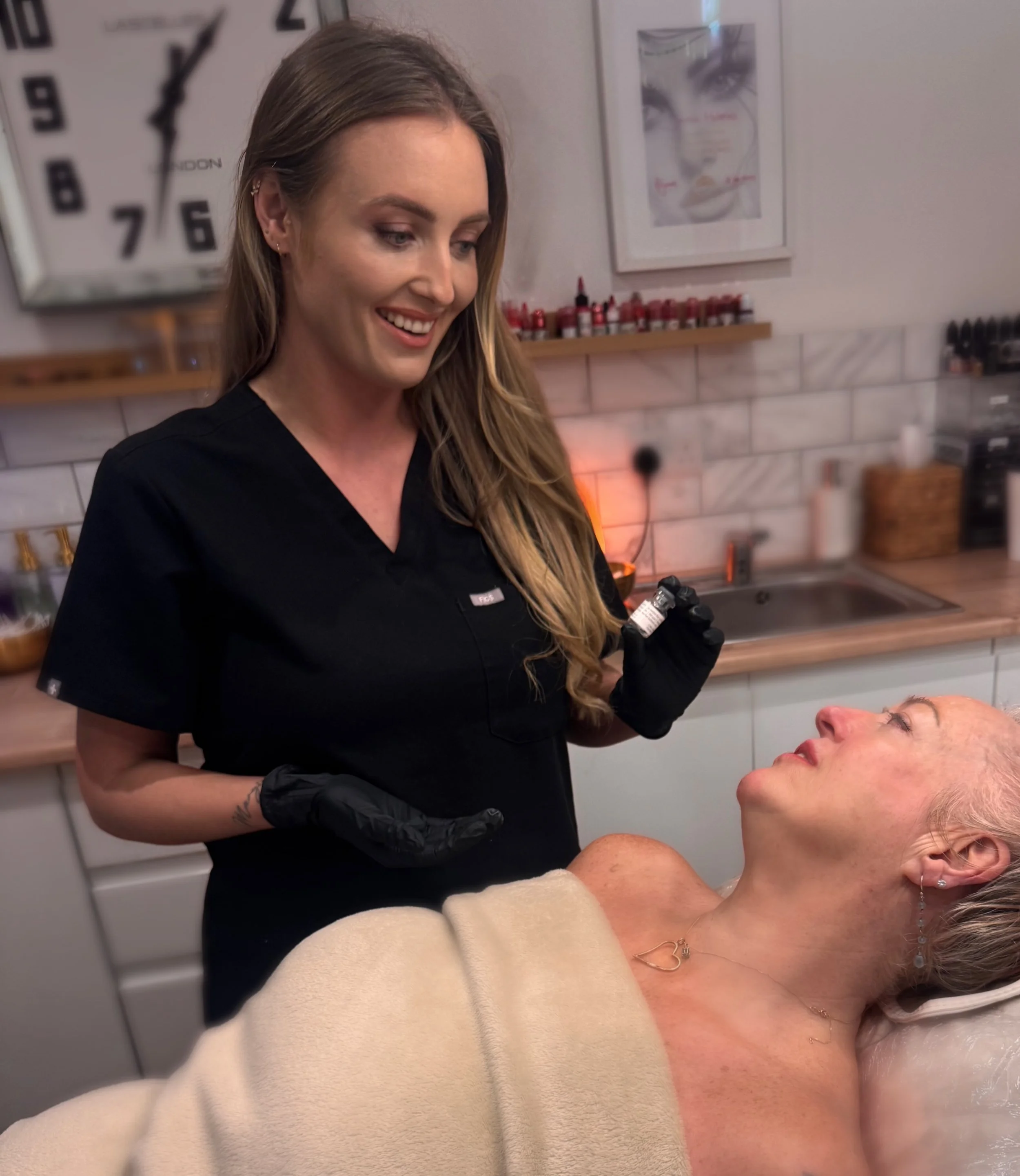 A healthcare professional in black scrubs and gloves smiling at an older woman lying on a treatment bed, ready to receive a cosmetic injection in a skincare clinic.