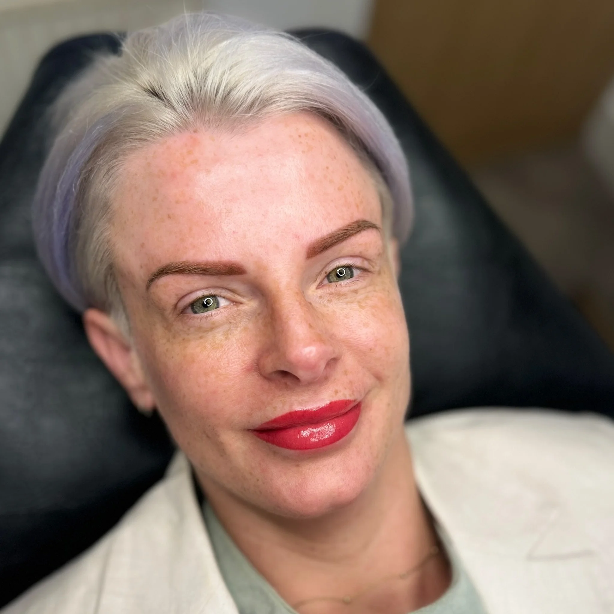 A woman with silver-gray hair lying back, smiling, with freckles, red lipstick, and light-colored clothing.
