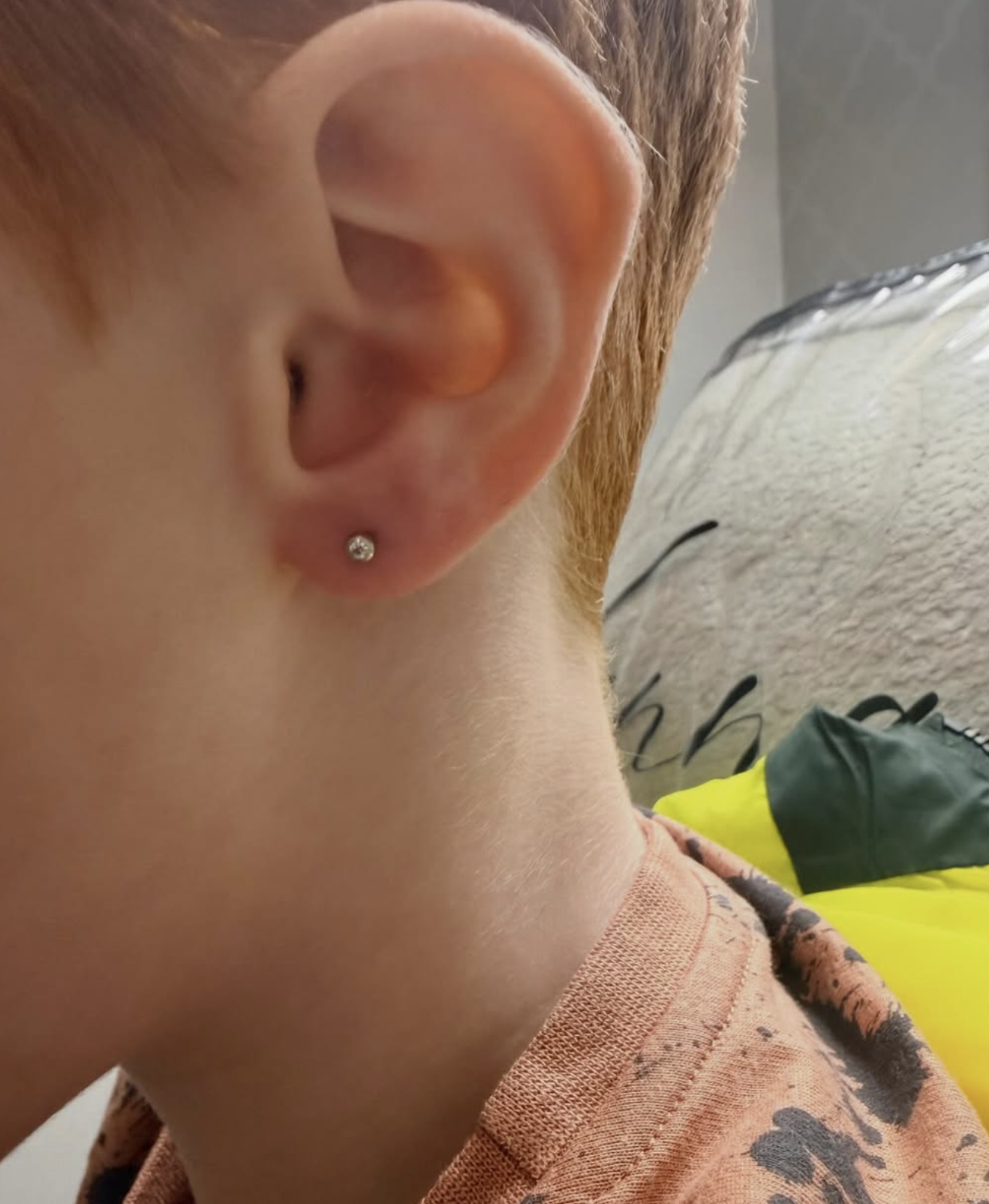 Close-up of a person's ear with a small diamond stud earring, part of their short red hair, and the side of their neck. They are wearing a peach-colored shirt with a dark pattern.