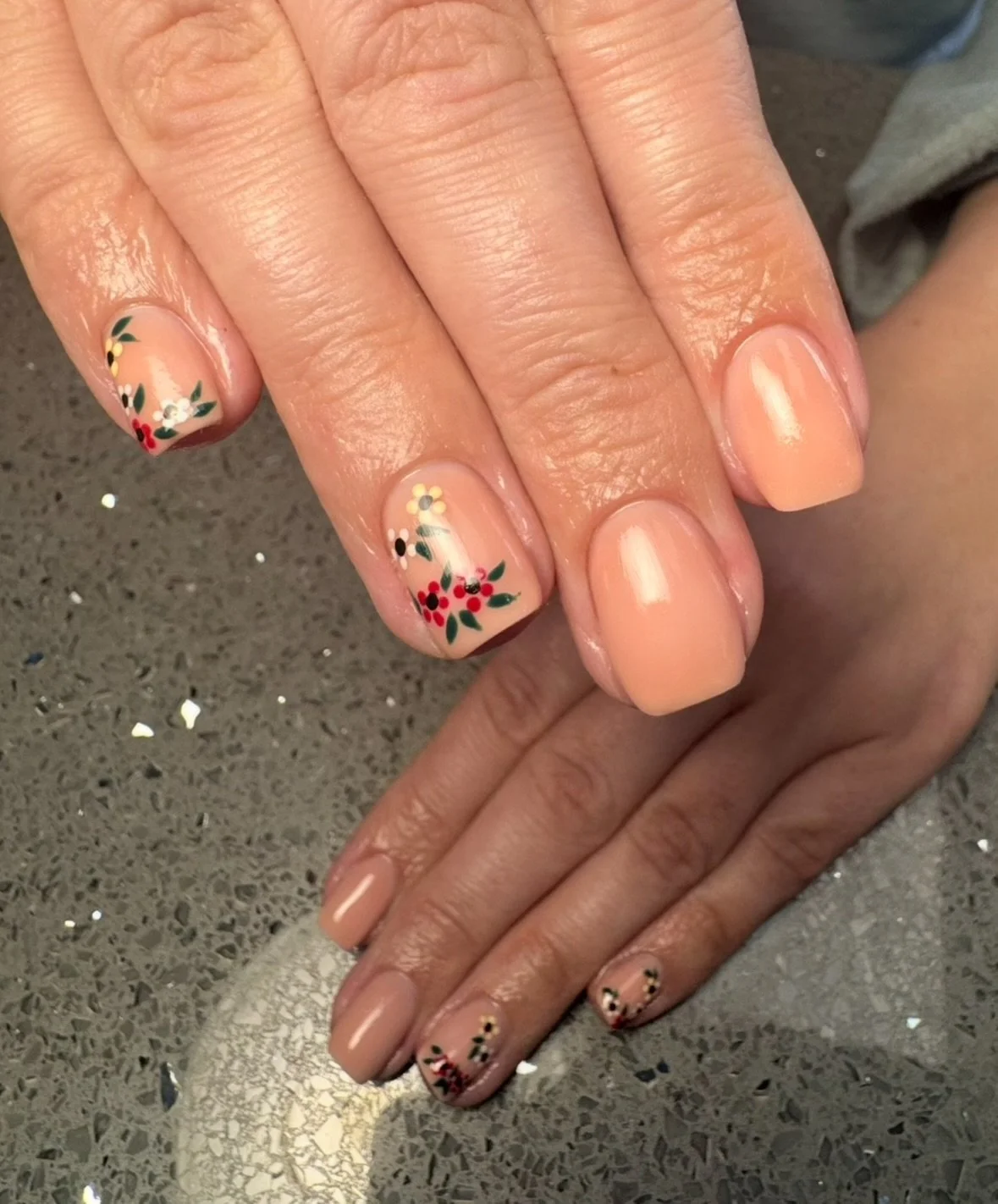 A close-up of a person's hands with natural-colored nails, decorated with small floral designs on the ring fingers and pinky fingers, featuring red, yellow, and black colors, on a speckled gray surface.