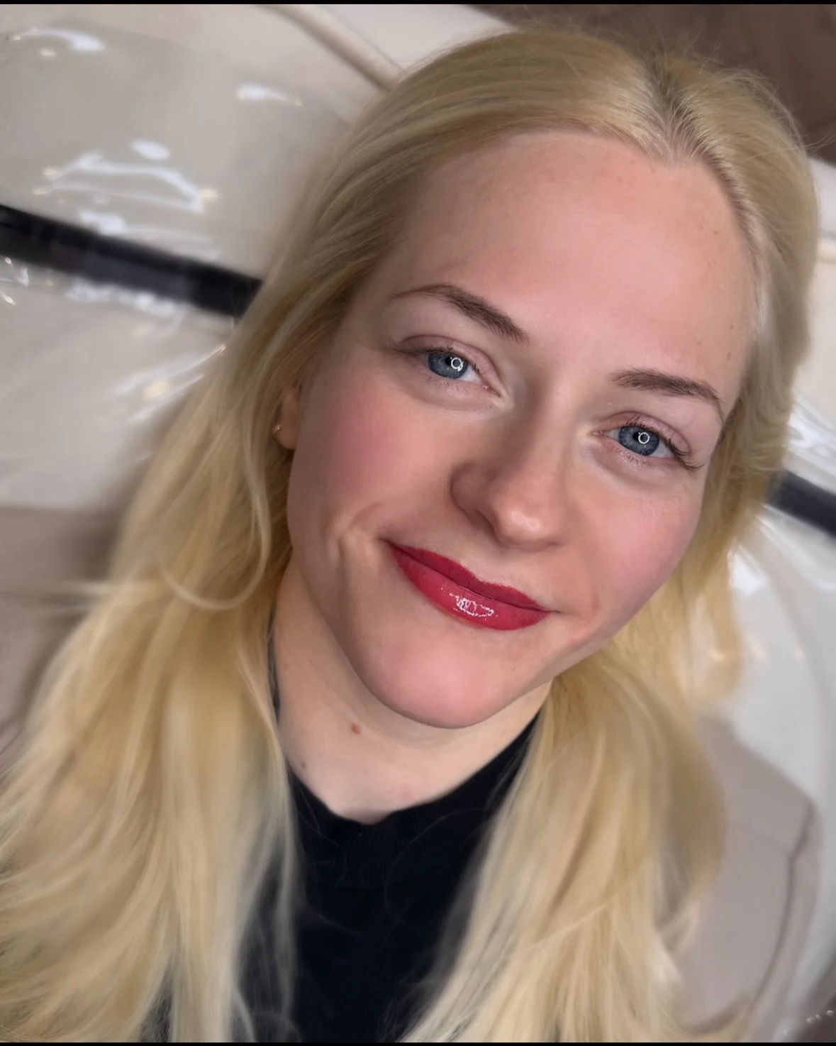 Close-up of a smiling woman with long blonde hair, bright blue eyes, wearing red lipstick, and a black top, lying down on a pillow.
