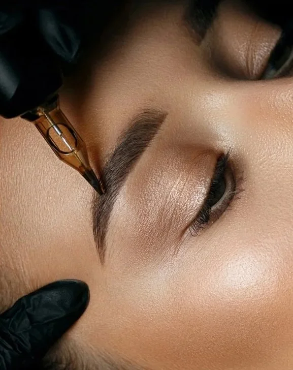A person receiving a cosmetic eyebrow tattooing procedure, with a gloved hand and tattooing device being used on the eyebrow.