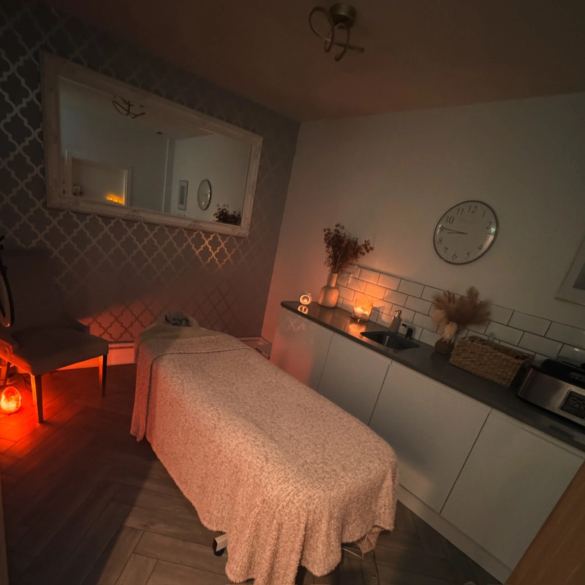 A massage table covered with a beige blanket in a dimly lit massage room. To the left, there is a gray chair and a glowing salt lamp on the floor. On the right, there is a countertop with lit candles, decorative vases with dried flowers, a clock, and various household items. A large mirror is on the wall, reflecting part of the room.