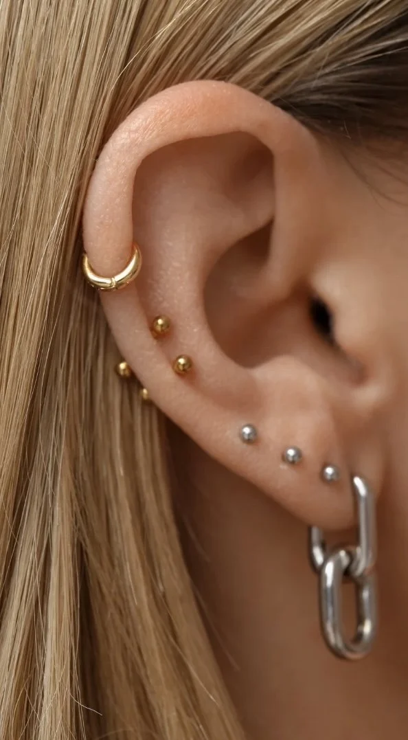 Close-up of a person's ear with multiple piercings, including a gold hoop earring on the upper cartilage, small gold and silver studs along the earlobe, and a large silver hoop earring.