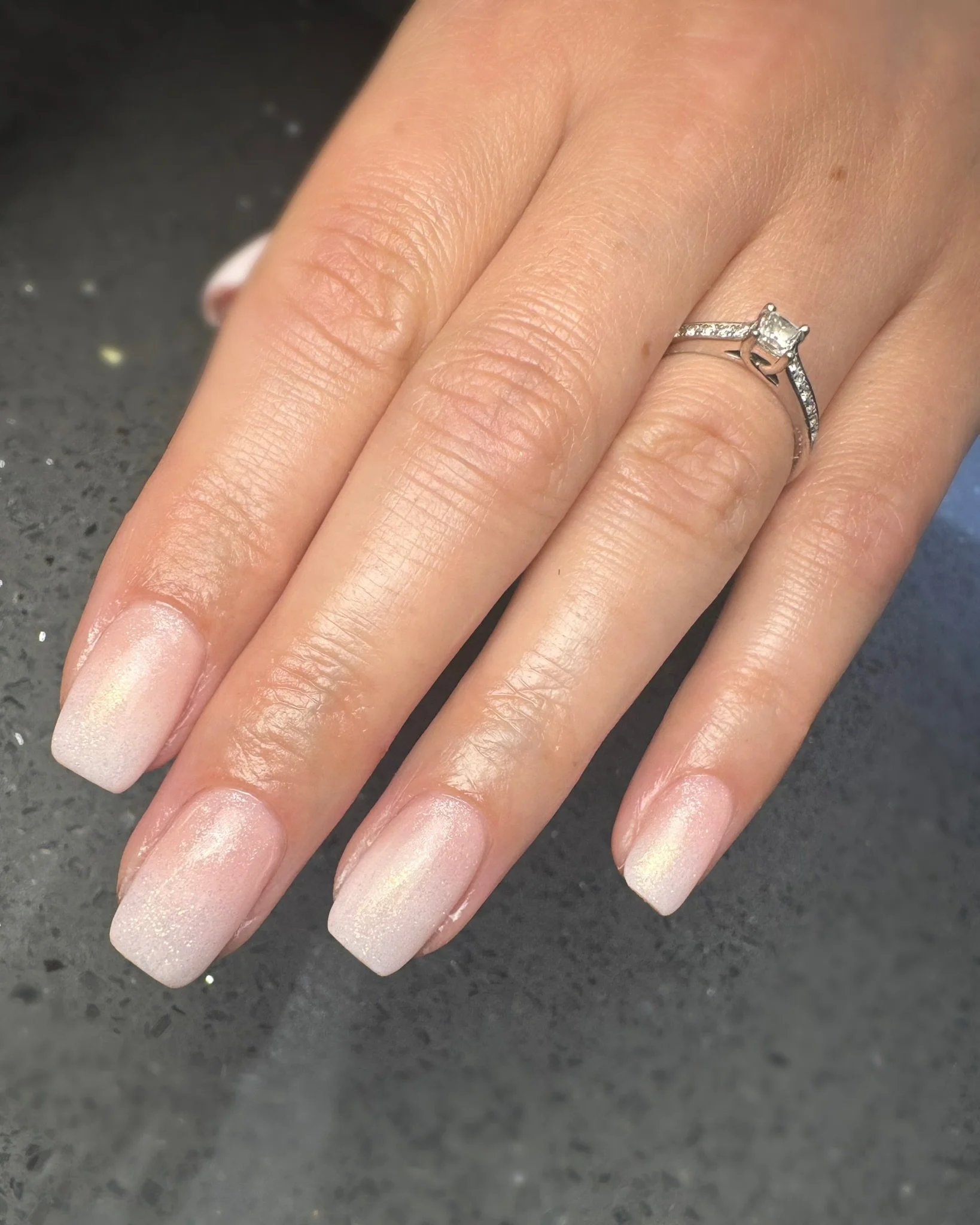 Close-up of a woman's hand with manicured nails and a diamond engagement ring on her finger, resting on a dark surface.