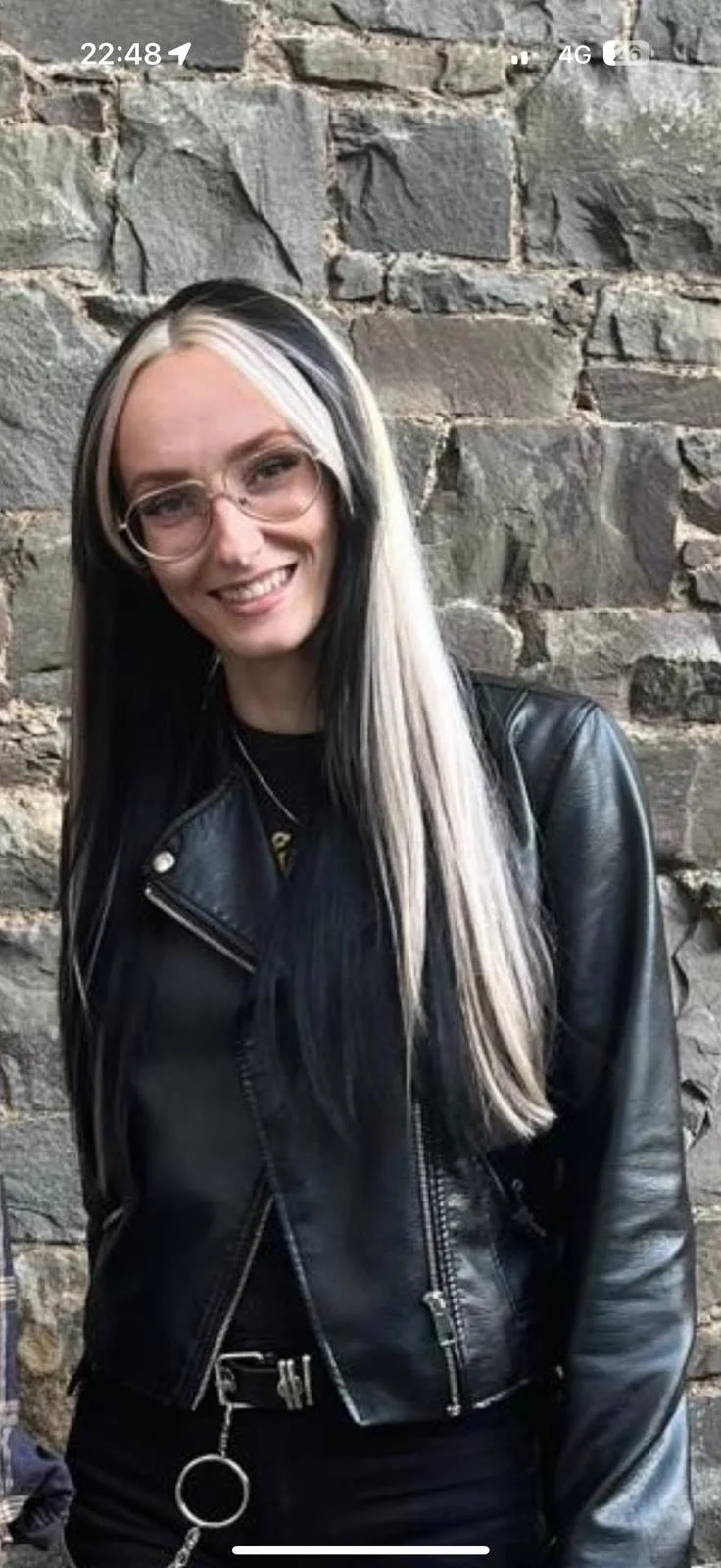 A woman with long black and platinum blonde hair, wearing glasses and a black leather jacket, smiling and standing against a stone wall.