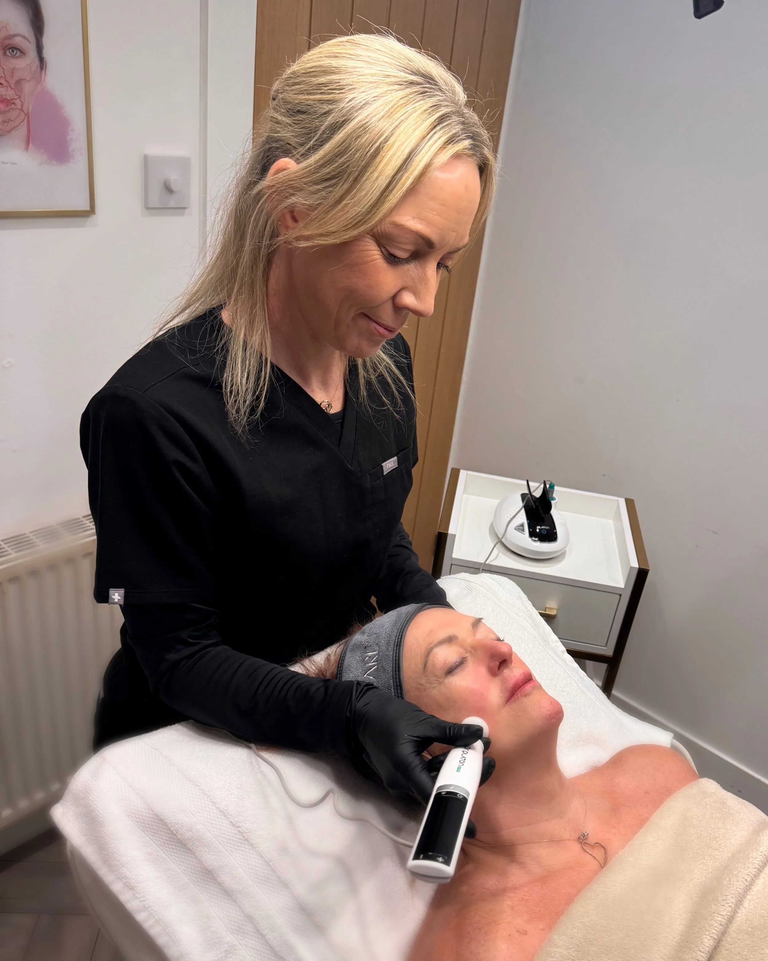 A woman in a black medical uniform performs a skin treatment on a woman lying on an examination bed. The practitioner holds a handheld device against the woman's cheek. The woman on the bed has a gray headband and appears relaxed.