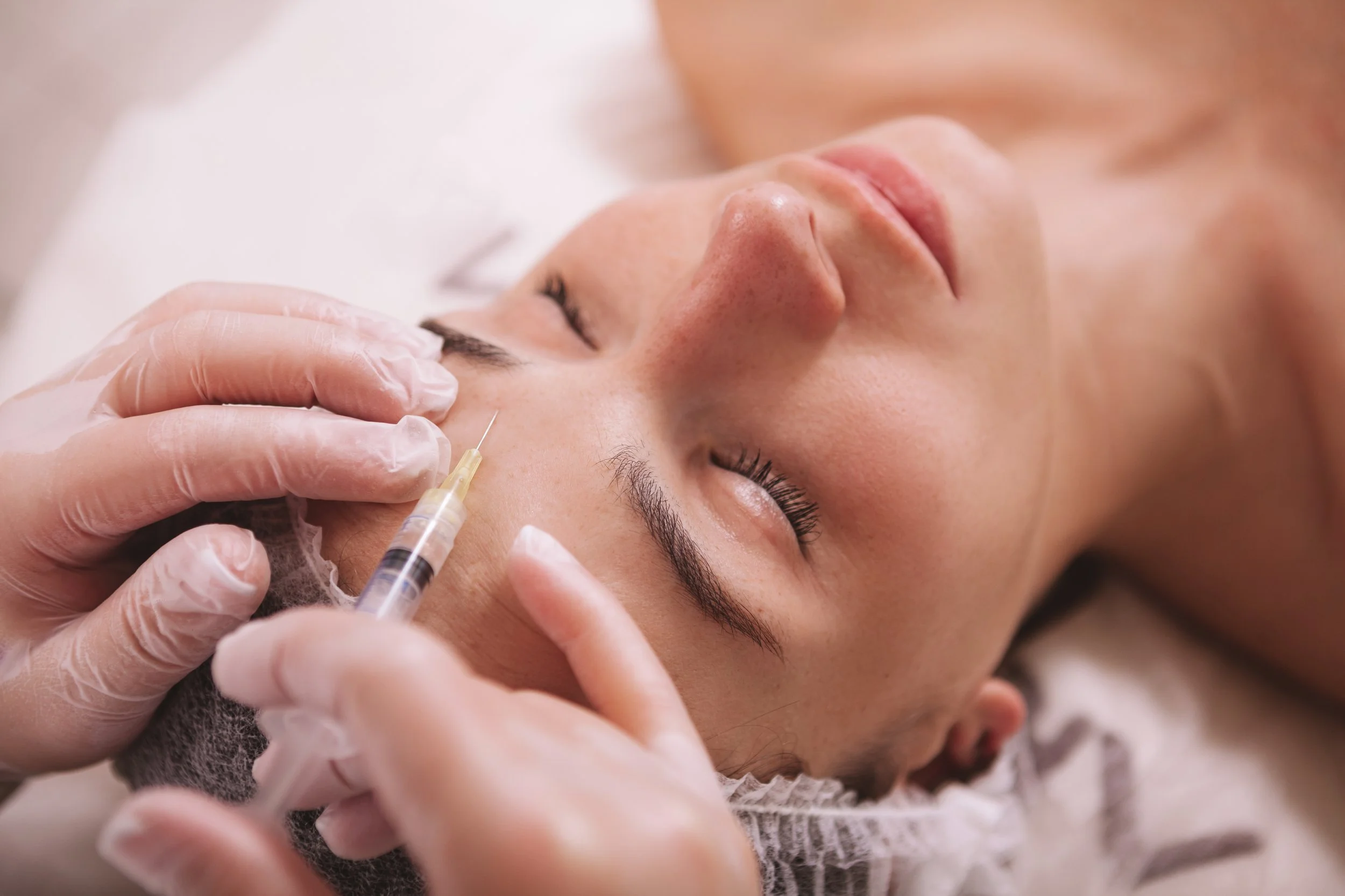 A woman lying down with eyes closed, receiving a cosmetic injection around her eyebrow area from a professional wearing gloves.
